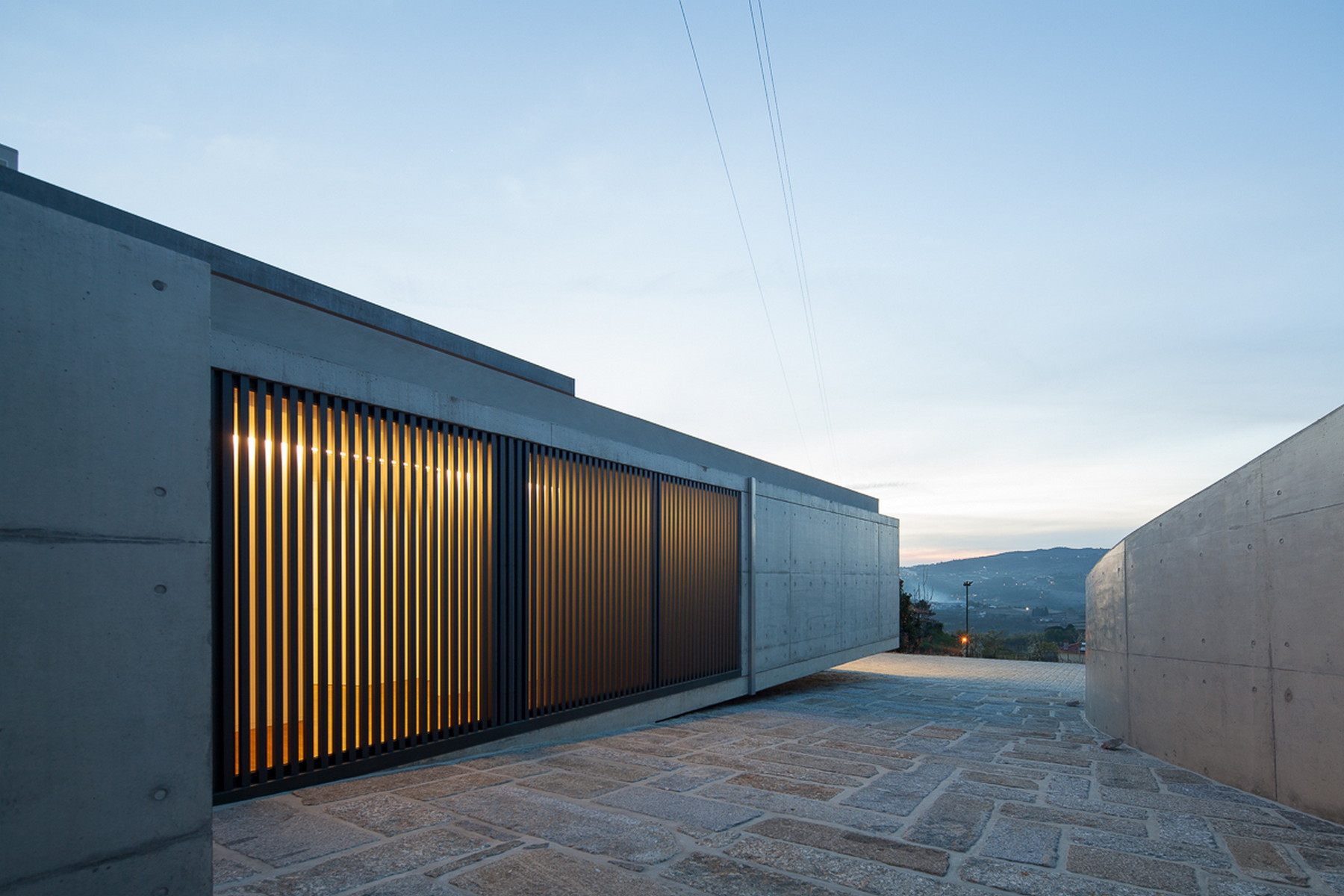 Board-formed concrete wall with a vertical slatted screen glowing amber, above a stone-paved entry court at dusk