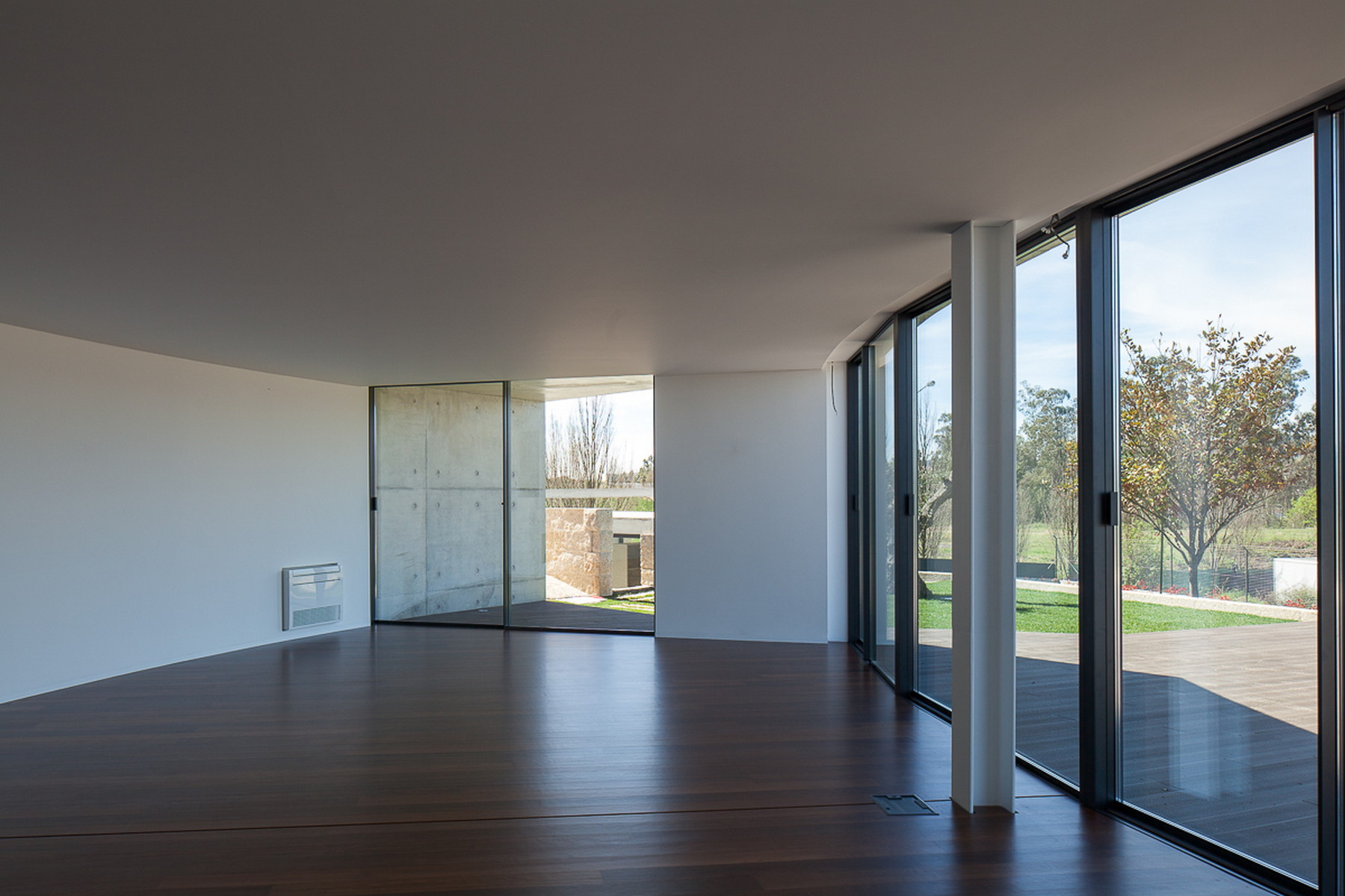 Empty living room with dark timber floor, a slim white column and sliding glass doors opening to a lawn and trees