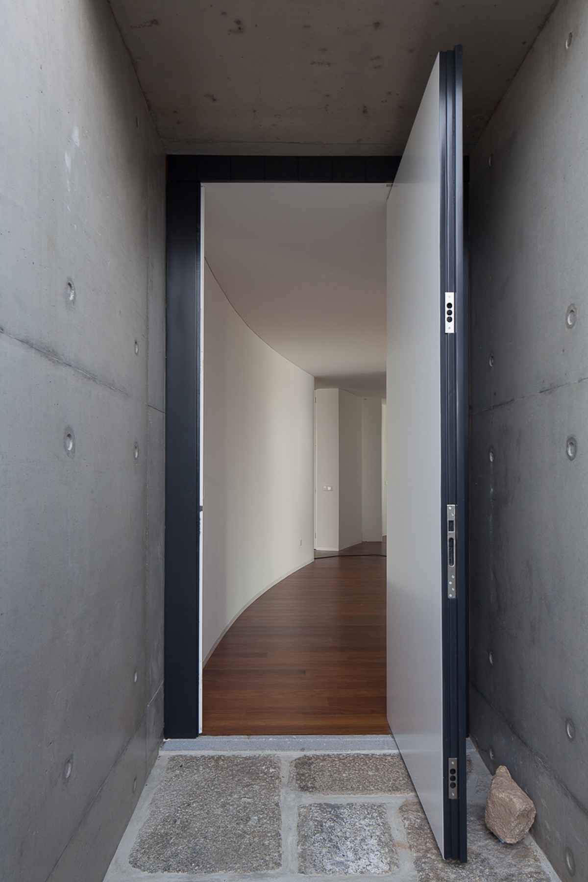 Pivoting front door held open between raw concrete walls, revealing a curved white corridor beyond a granite threshold
