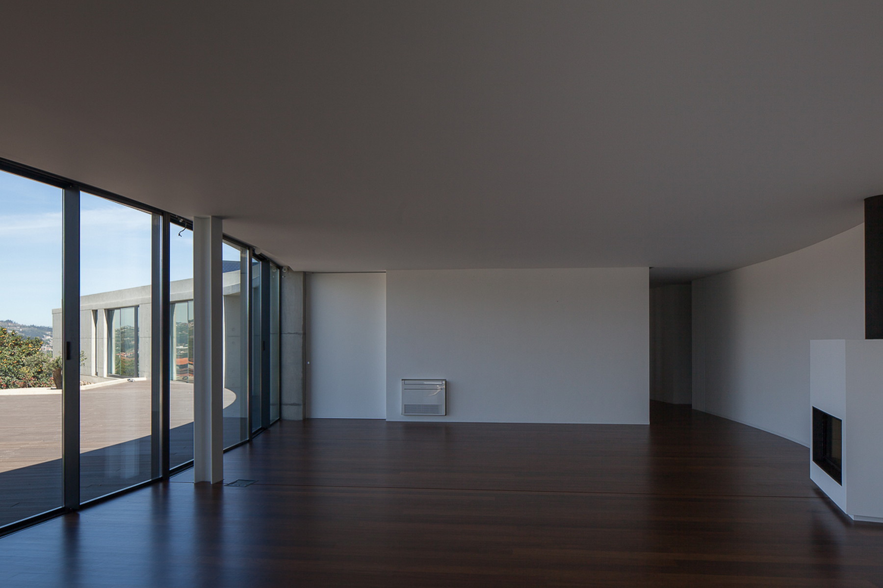 Open living space with a curved white wall, built-in fireplace and floor-to-ceiling glazing facing a timber terrace in daylight