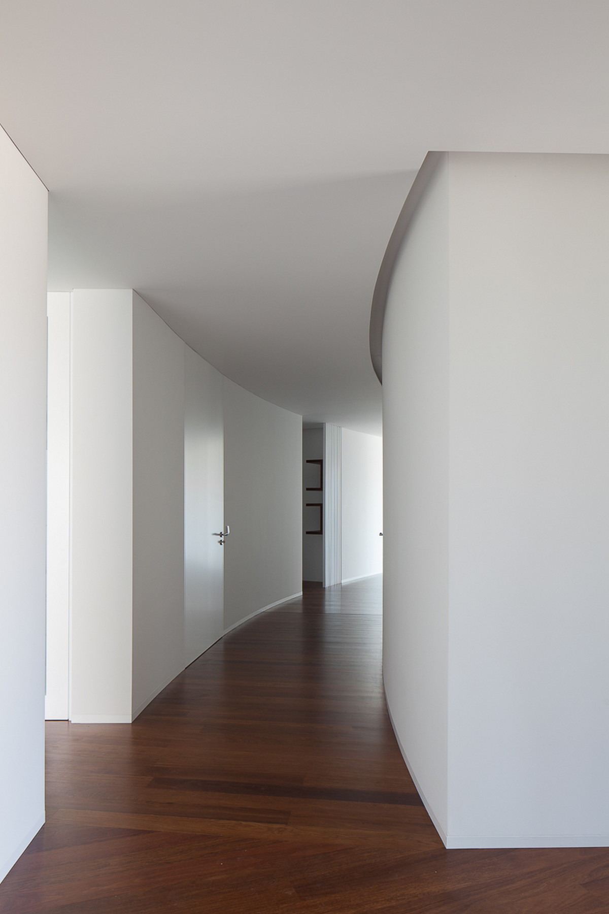 Curved white corridor with flush doors and a dark timber floor, narrowing toward framed pictures in the distance