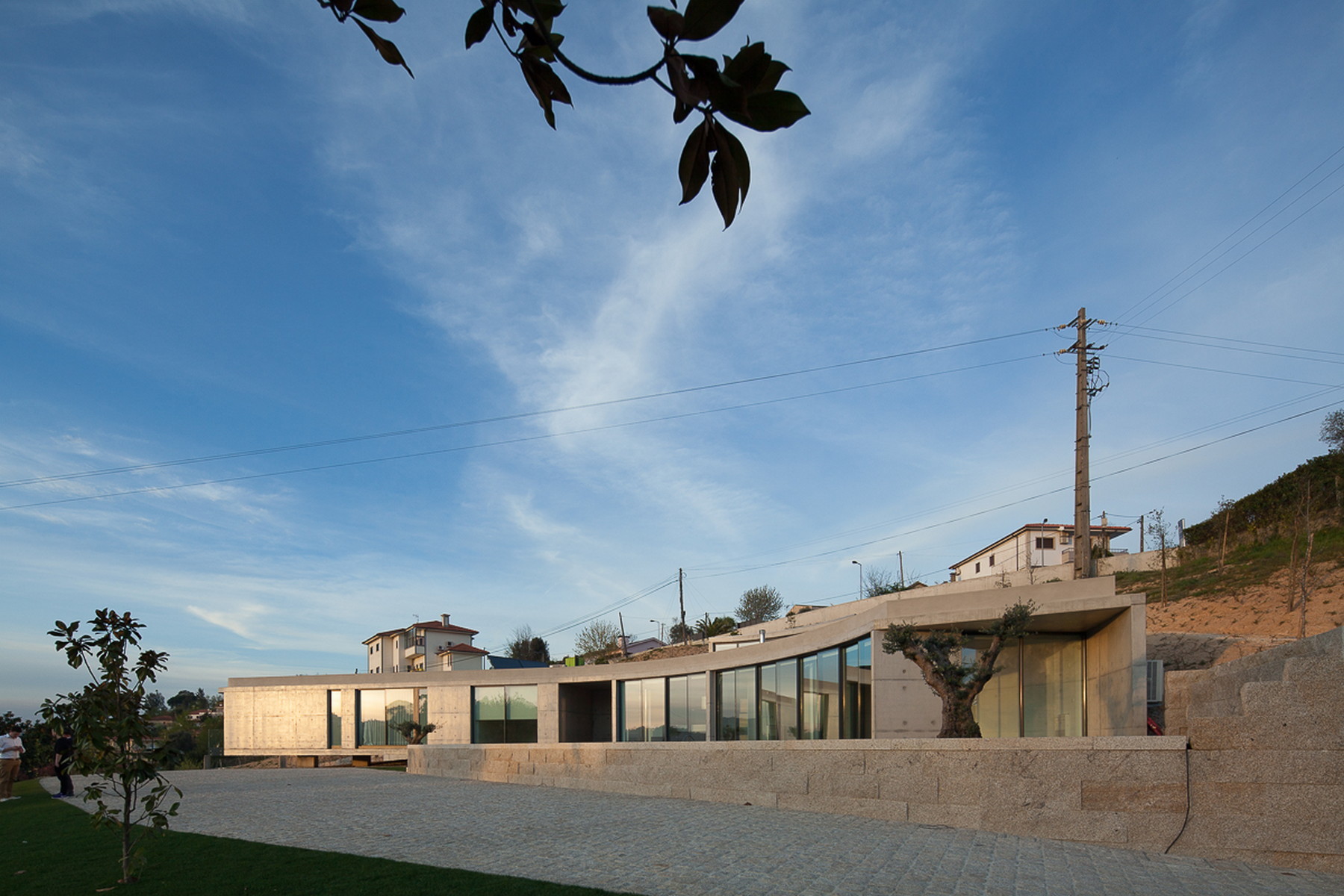 Low curved house with floor-to-ceiling glazing behind a granite retaining wall and cobbled forecourt under a wispy blue sky