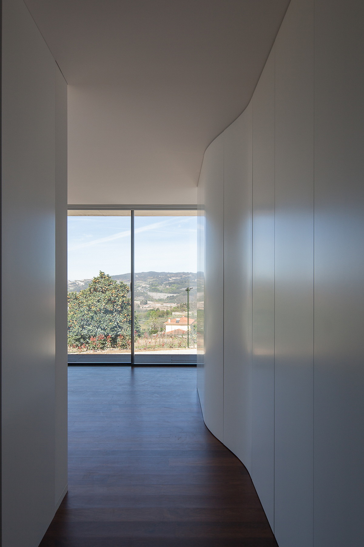 Curved white corridor wall leading to a full-height glazed opening framing a hillside view in daylight