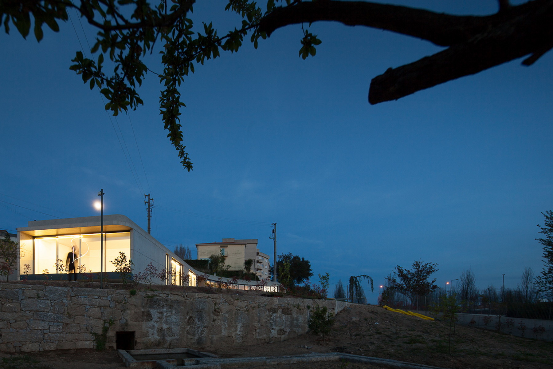 Glowing glazed end of the house seen from a stone-walled slope at dusk, with a figure inside and a branch overhead