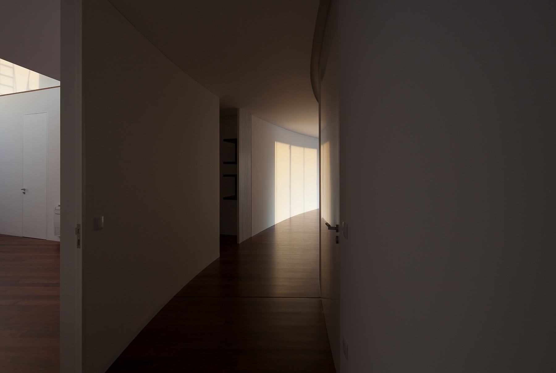 Dim curving hallway with white walls and dark timber floor, lit by a warm glow from the far window