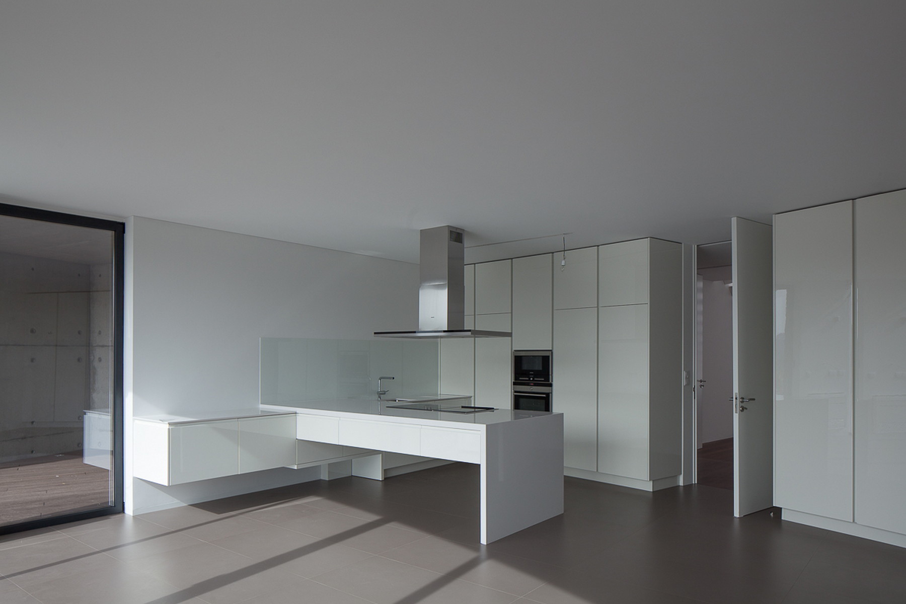 White kitchen with a floating counter, tall cabinets and extractor hood, with sunlight raking across the grey floor