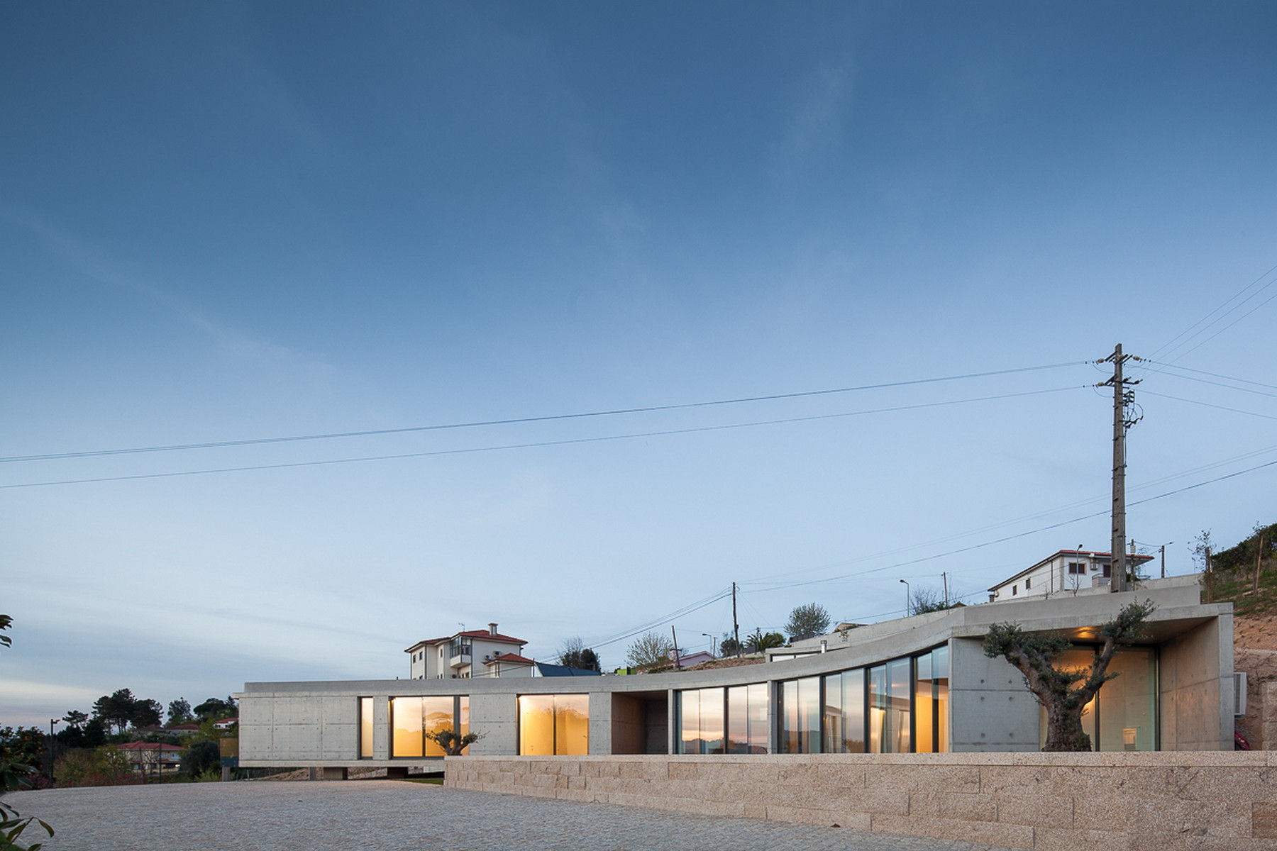 Long concrete house with glazed openings glowing at dusk, a granite retaining wall and an olive tree in the foreground