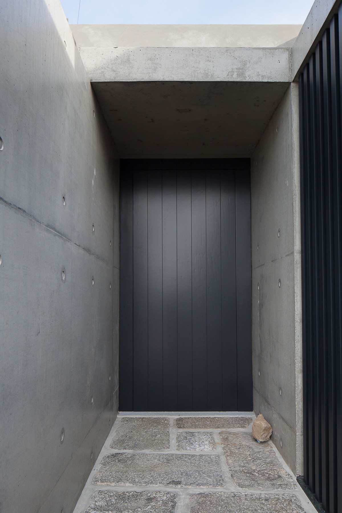 Recessed entrance between board-marked concrete walls, with a black vertical-slat door and a granite paved threshold under pale sky
