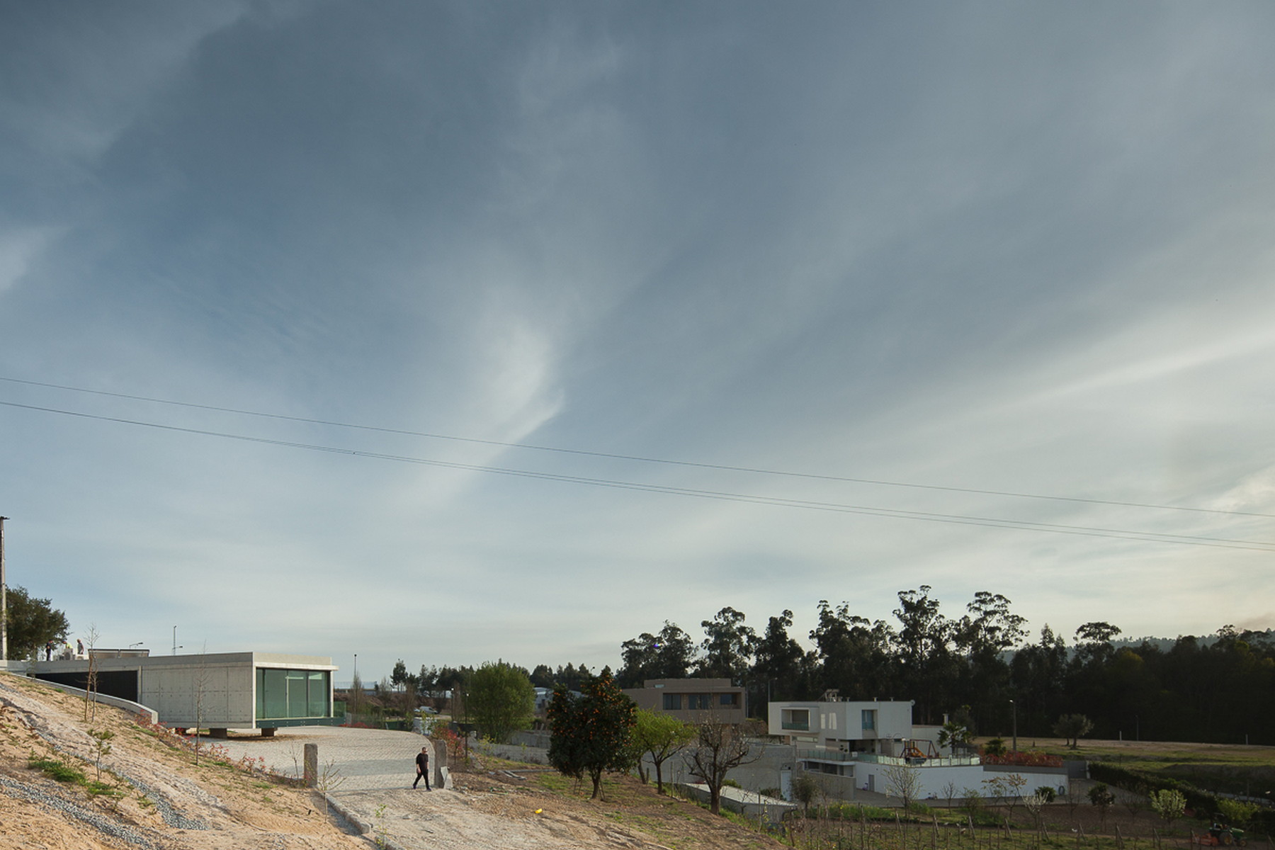 Distant view of the low concrete volume on a hillside, with a lone walking figure and wispy clouds overhead