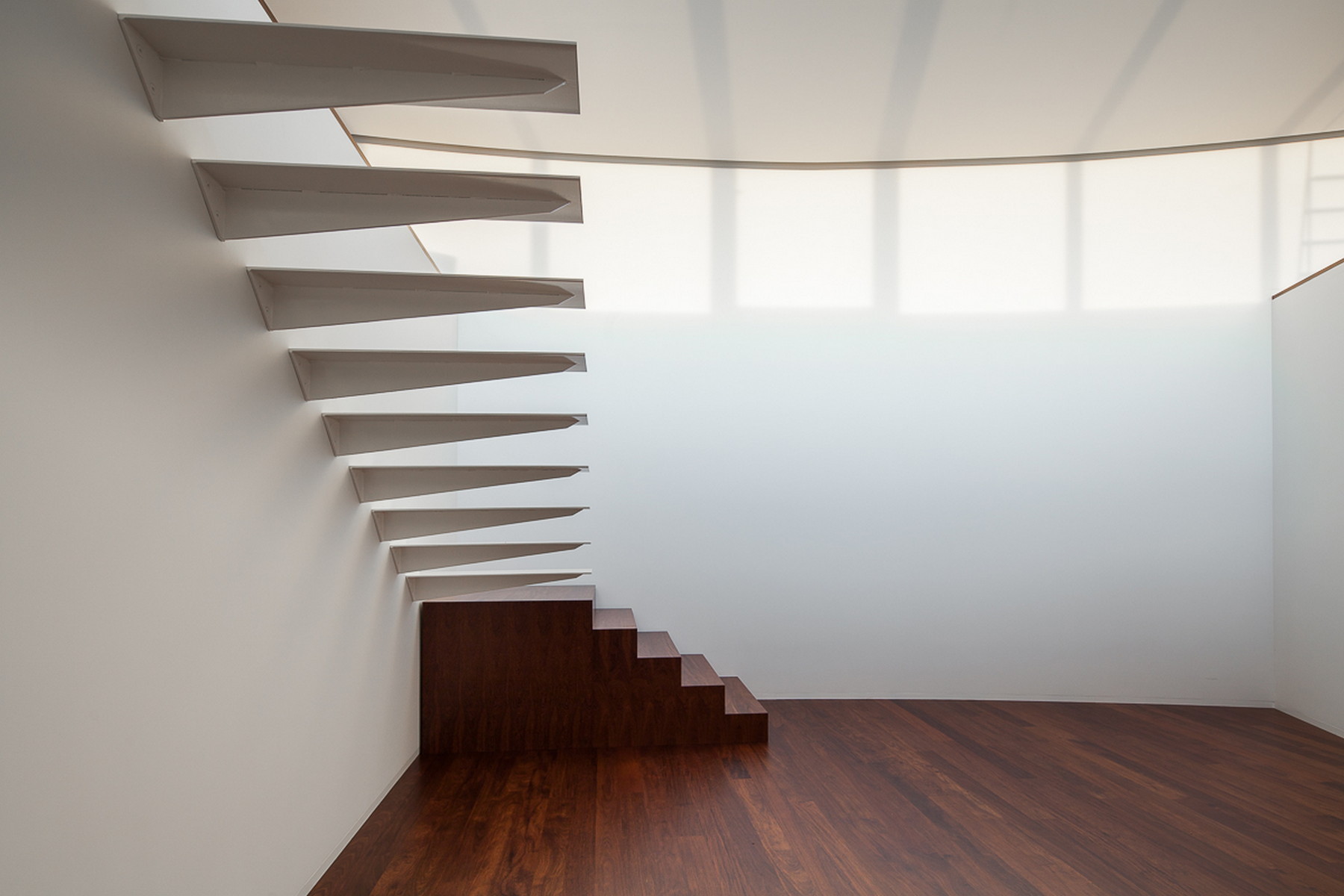 Cantilevered white stair treads projecting from a wall above a dark timber stair base and wood floor, lit by a soft translucent window