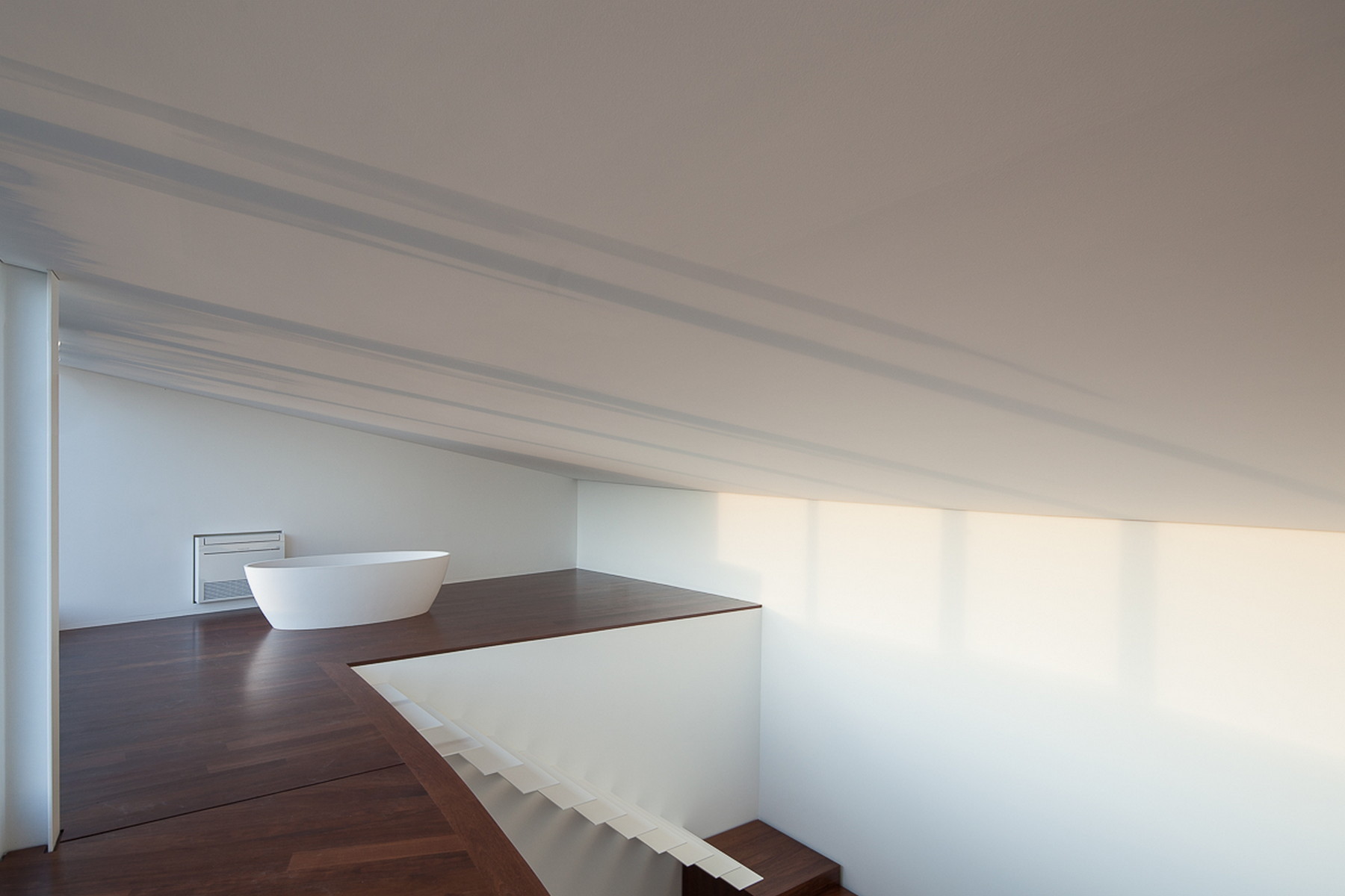 Upper floor under a sloped white ceiling with a freestanding bathtub on a timber platform beside the stair opening