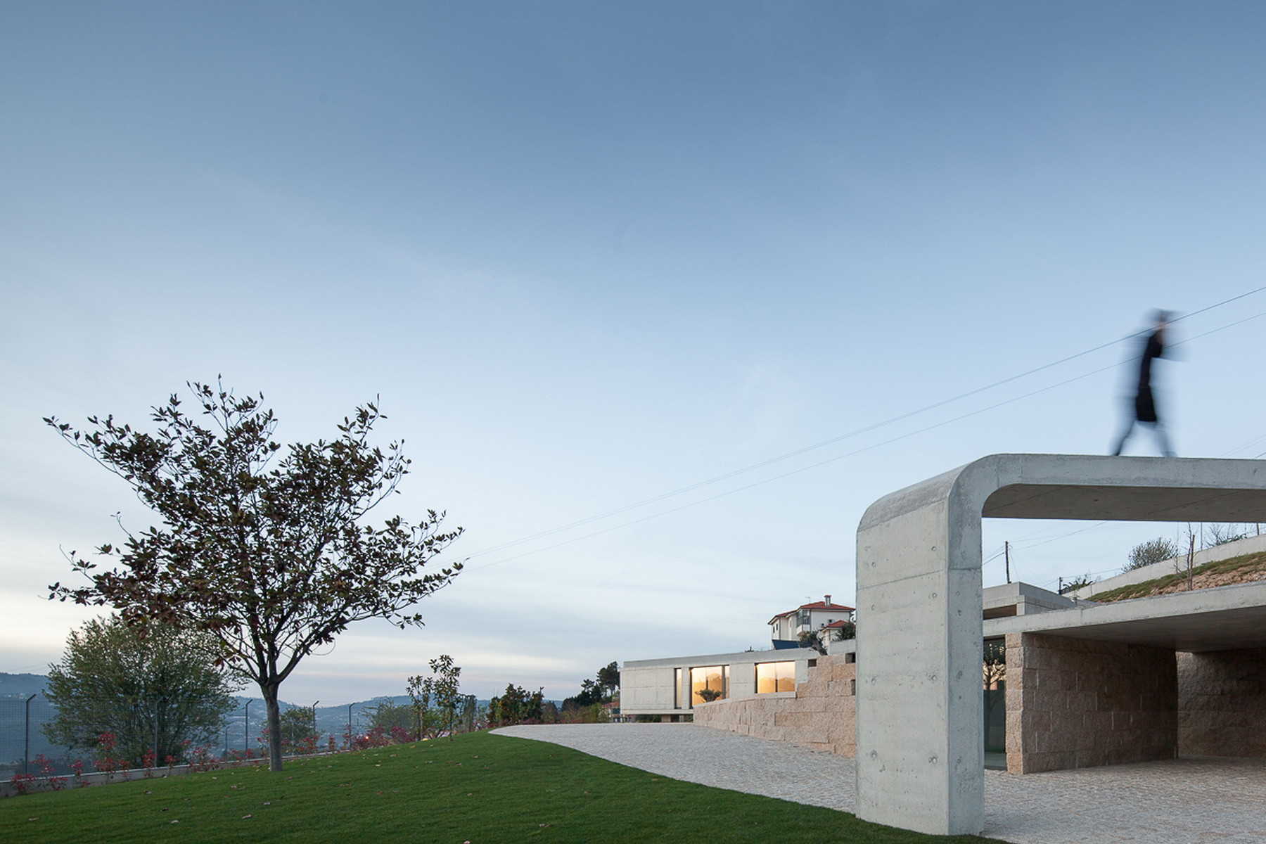 Concrete portal frame with a blurred figure walking across its roof, beside a granite wall, lawn and a bare tree at dusk