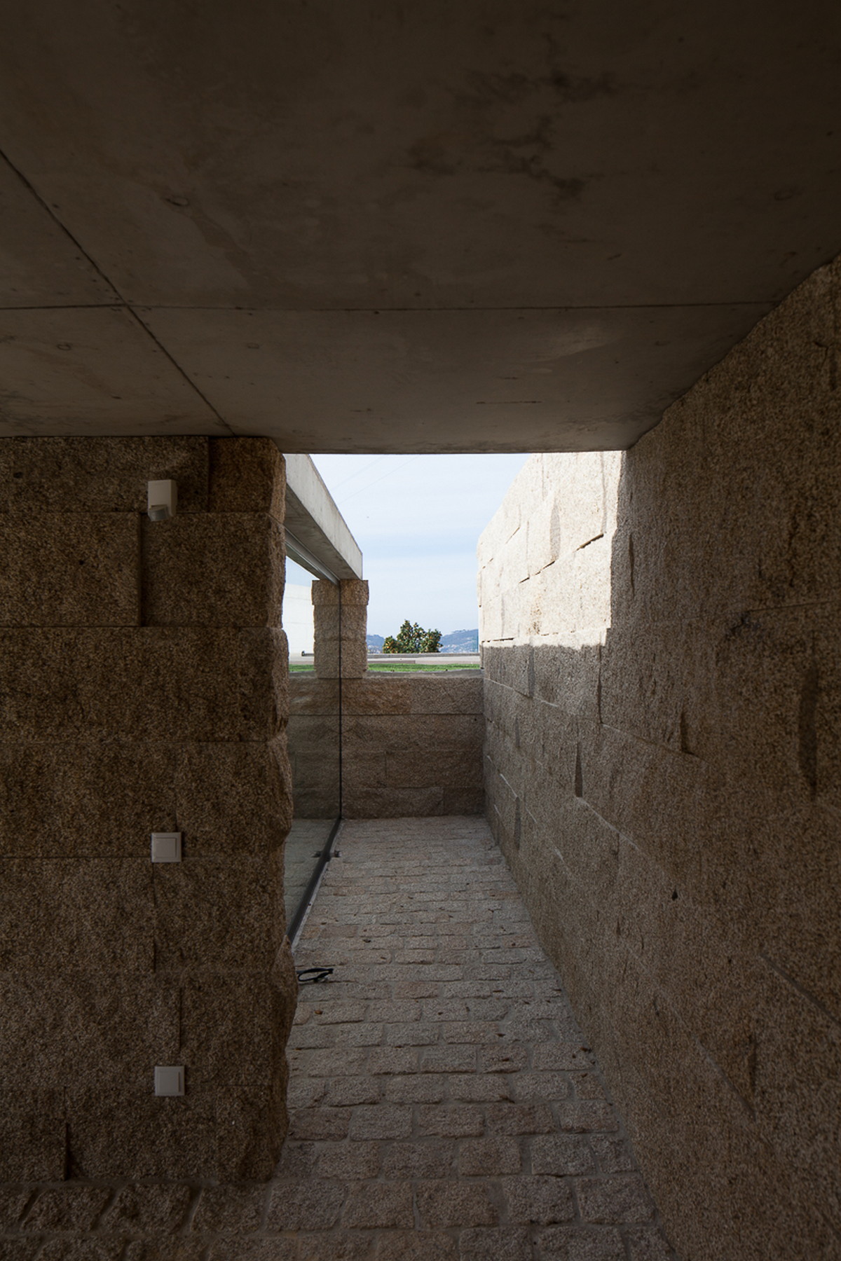 Narrow passage between rough-cut granite block walls under a concrete slab, framing a distant green lawn and hills in daylight