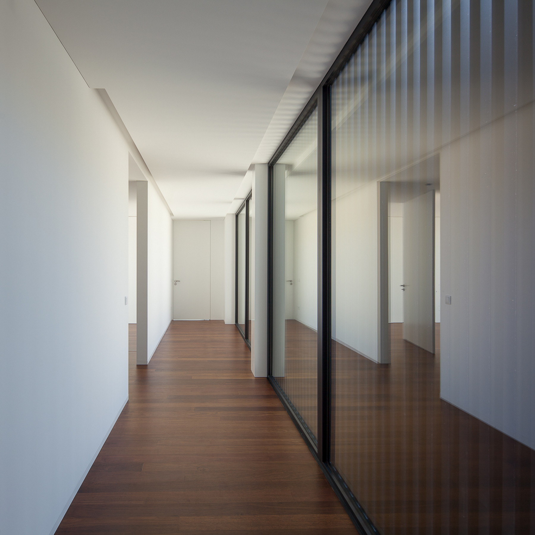 Long corridor with white walls, dark timber floor and a black-framed glass partition reflecting the doors in soft daylight