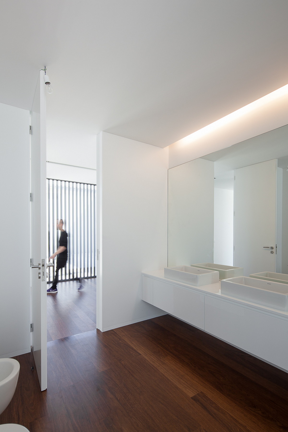 Bathroom with a floating white vanity, two basins and a long mirror, while a blurred person walks past the open door