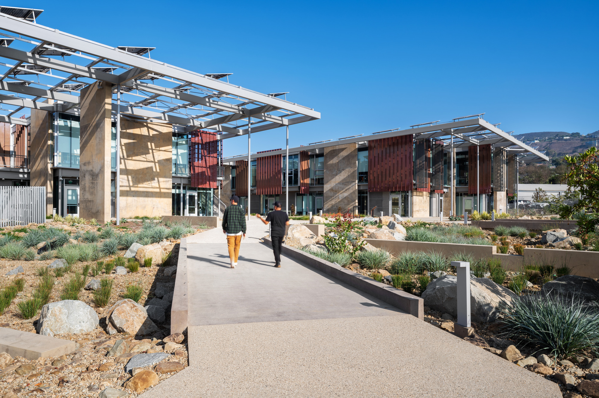 Concrete pathway through desert landscaping leading to glass and corrugated metal buildings under solar canopies