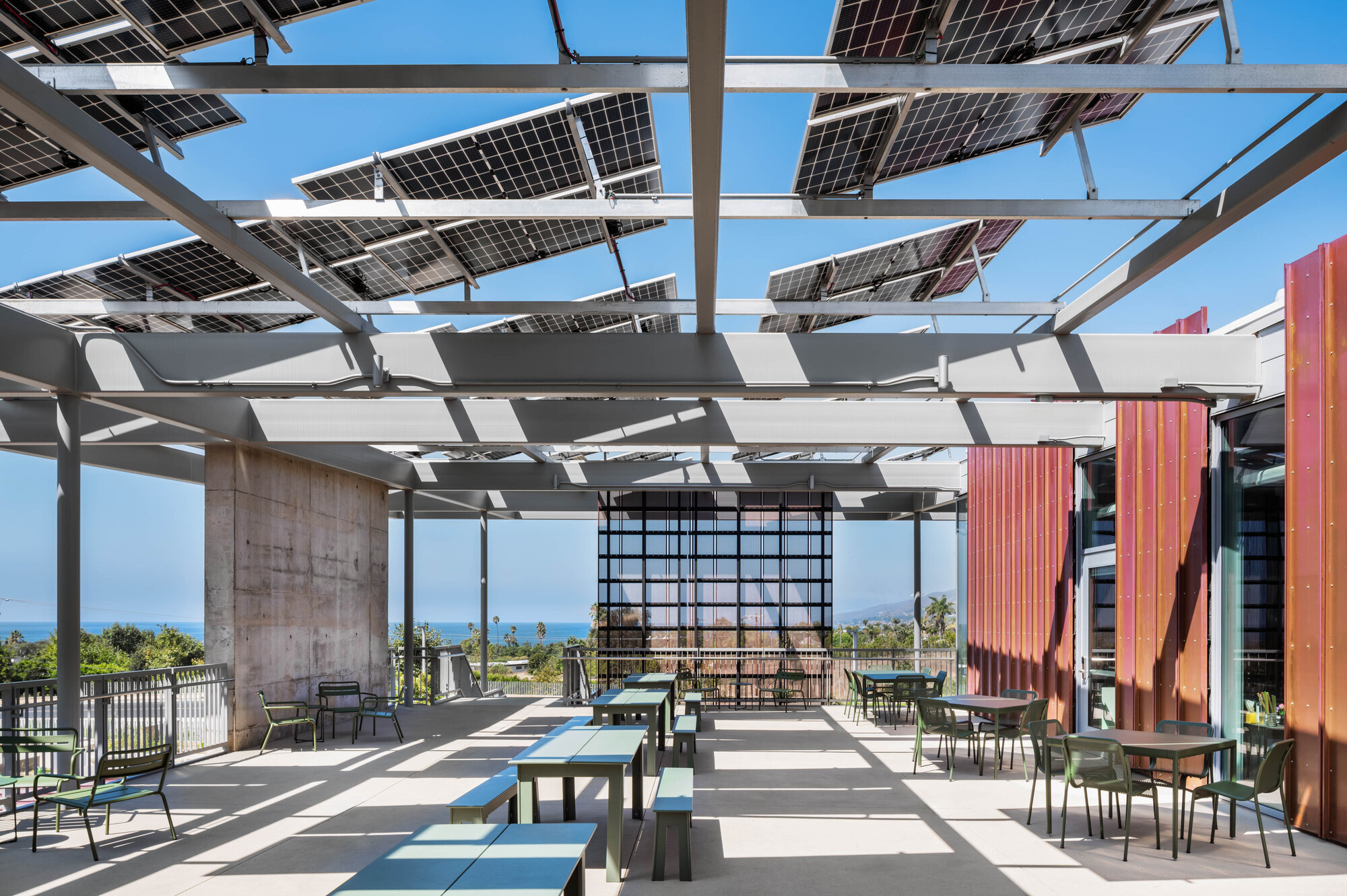 Rooftop terrace with solar panel canopy casting shadows over tables and chairs with ocean views beyond