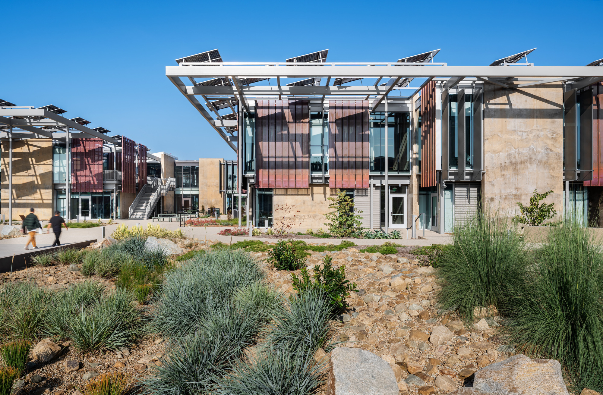 Facade with perforated red metal screens and rammed earth walls flanking a landscaped courtyard with native grasses