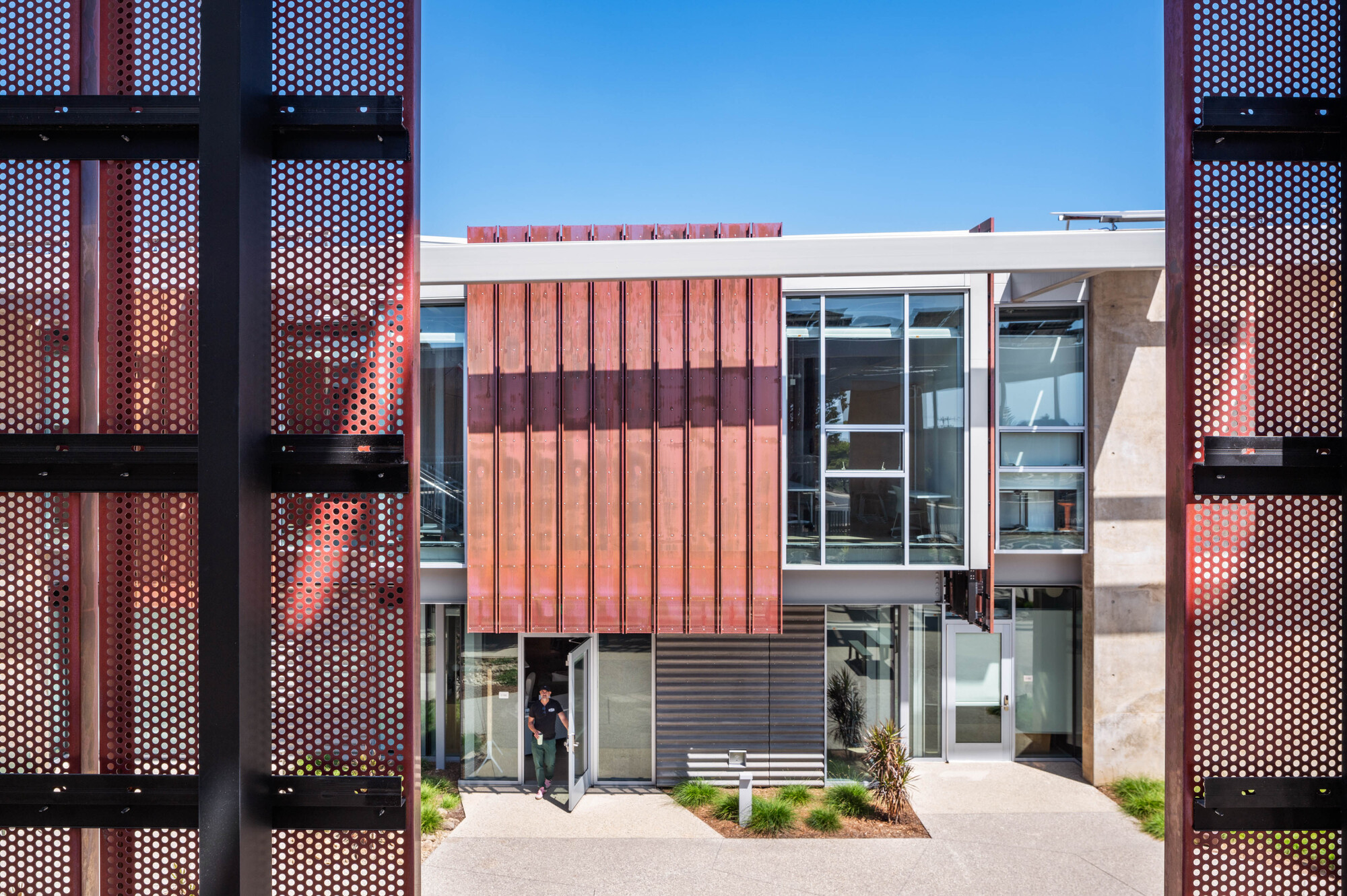View through perforated metal screens framing a two-story facade with vertical red panels and glazing