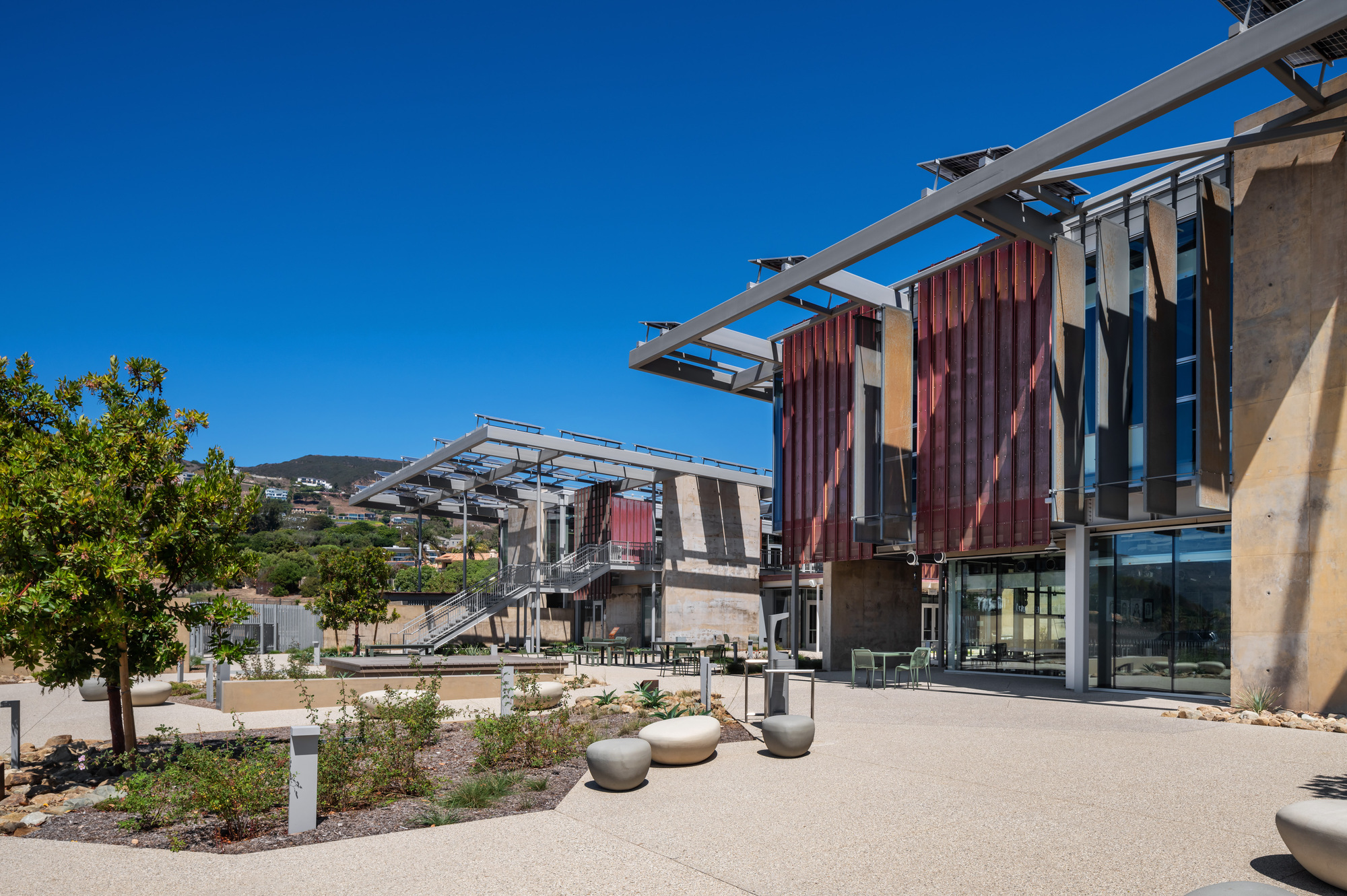 Campus courtyard with young trees and seating beneath solar canopy structures on a clear day