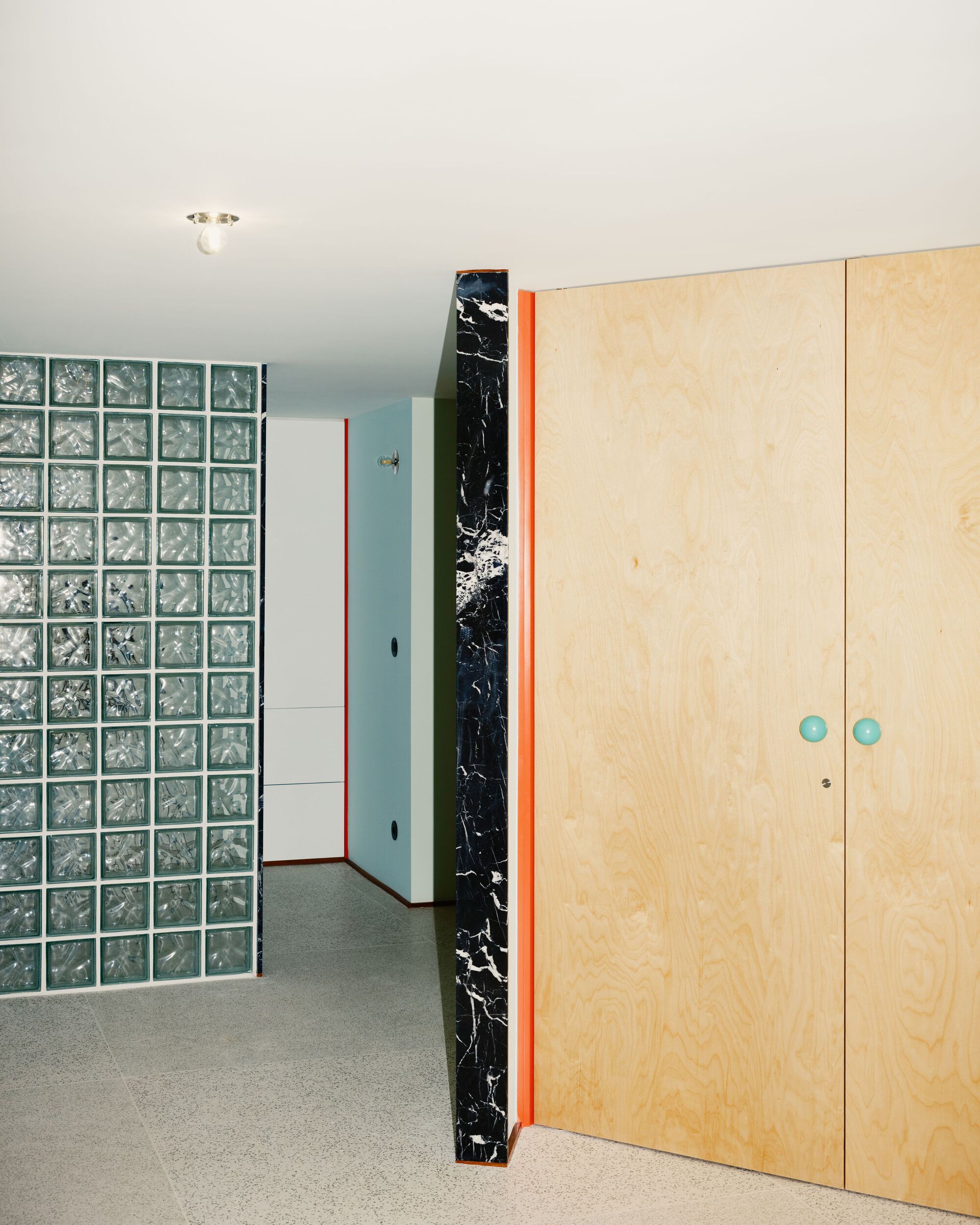 Light plywood wardrobe with blue knobs beside a glass block wall and black marble column