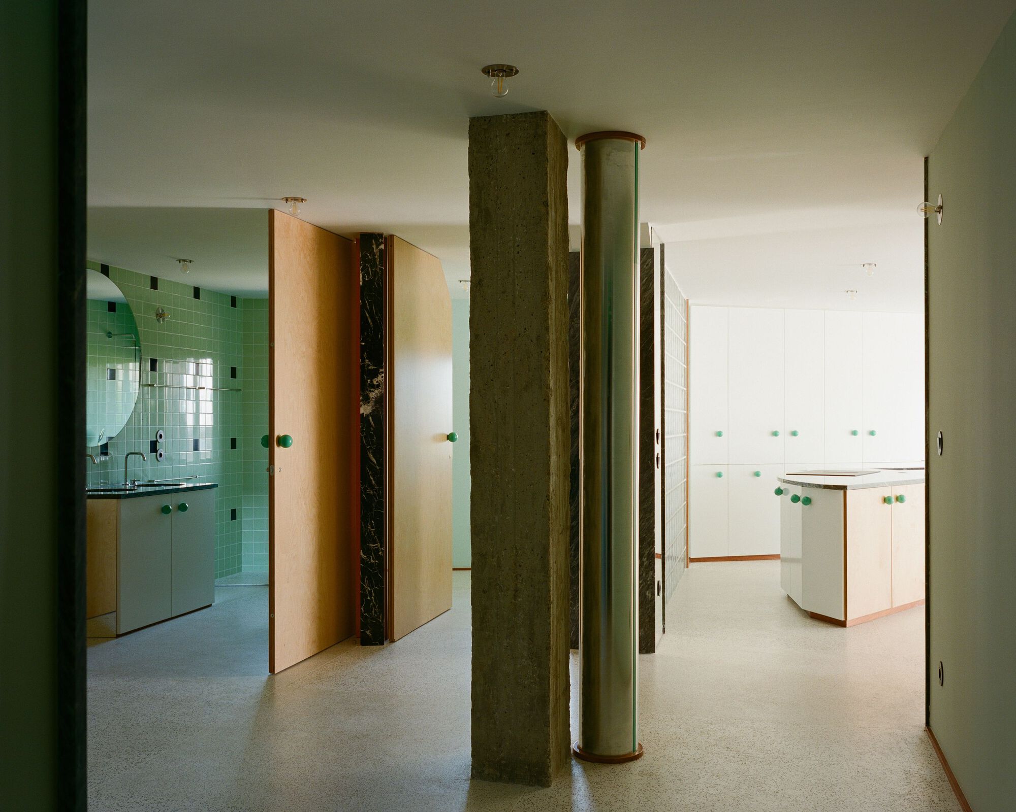 Open-plan interior with green glass block bathroom wall, cylindrical columns, and terrazzo flooring