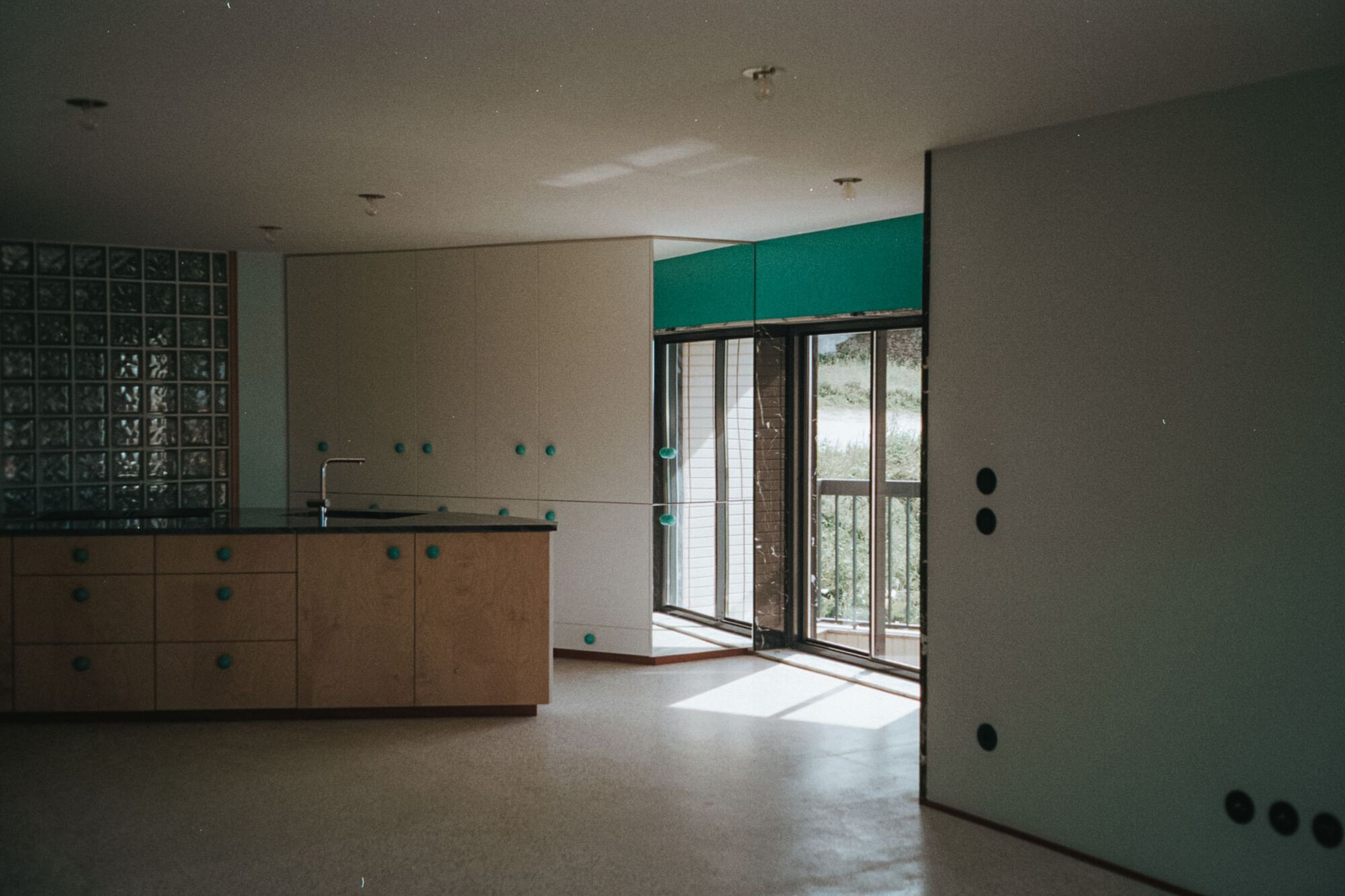 Kitchen with light plywood cabinetry, teal upper panels, and glazed balcony door with exterior blinds