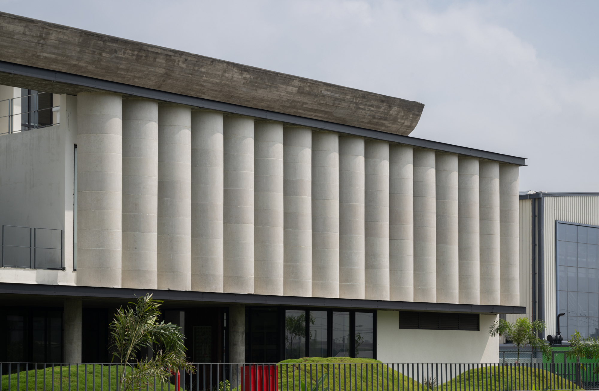 Oblique view of the ribbed concrete volume with planted foreground and adjacent industrial shed