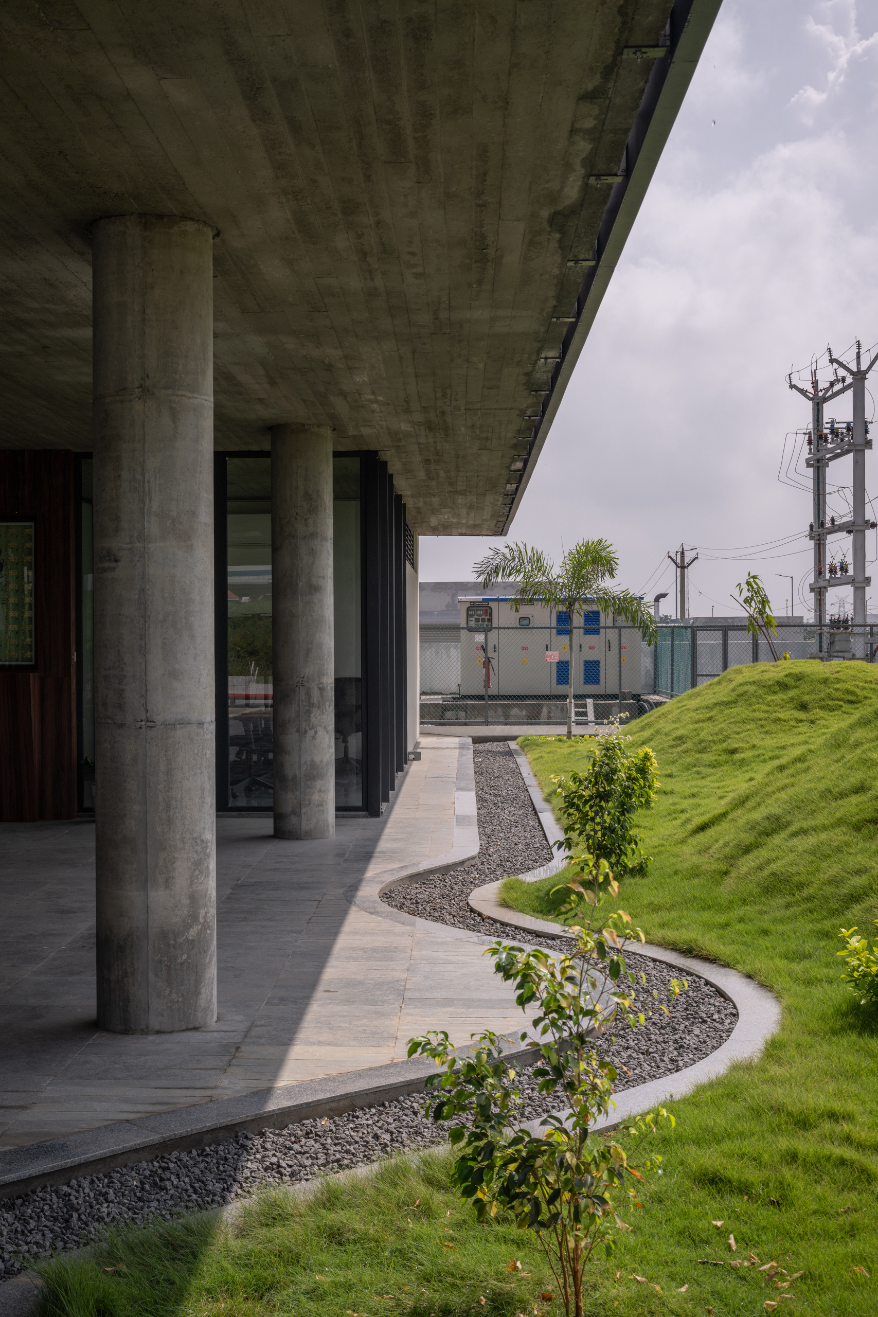 Covered walkway under board-formed concrete soffit with cylindrical columns and curved planting beds beyond