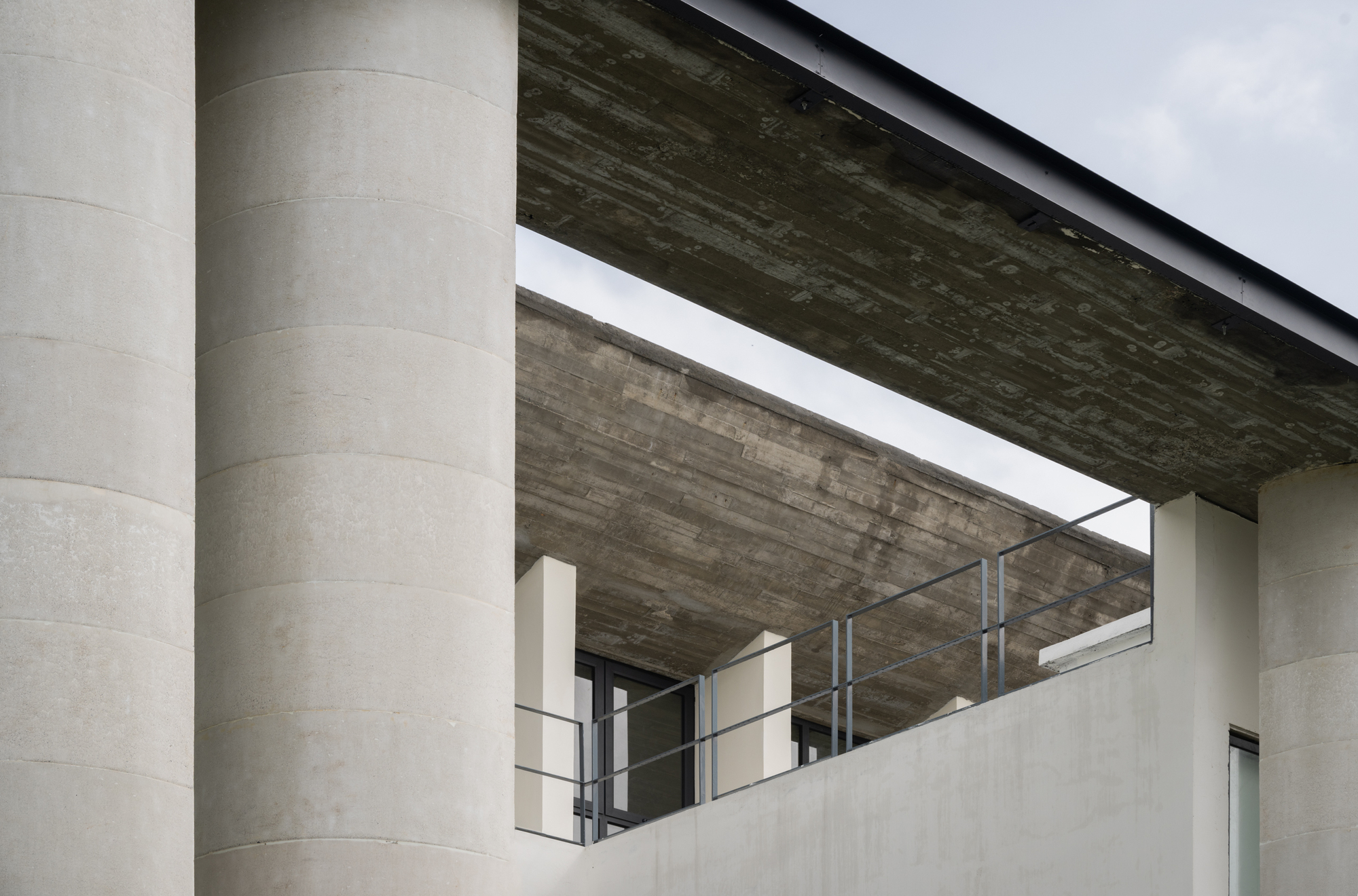 View beneath the cantilevered concrete roof showing cylindrical columns and metal railings under an overcast sky