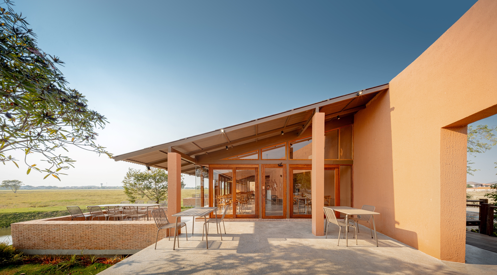 Covered terrace with timber columns and glazed dining hall opening to planted lawn and agricultural fields
