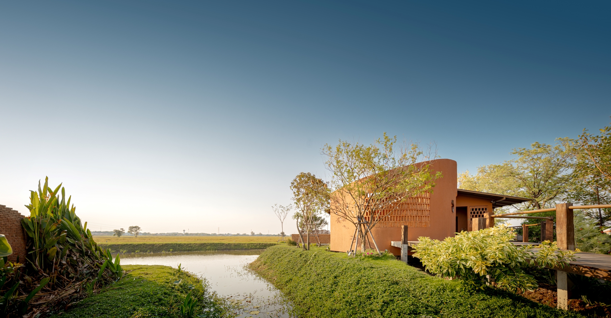 Perforated cylindrical tower and timber pavilion beside a pond with sloped planted banks at dusk