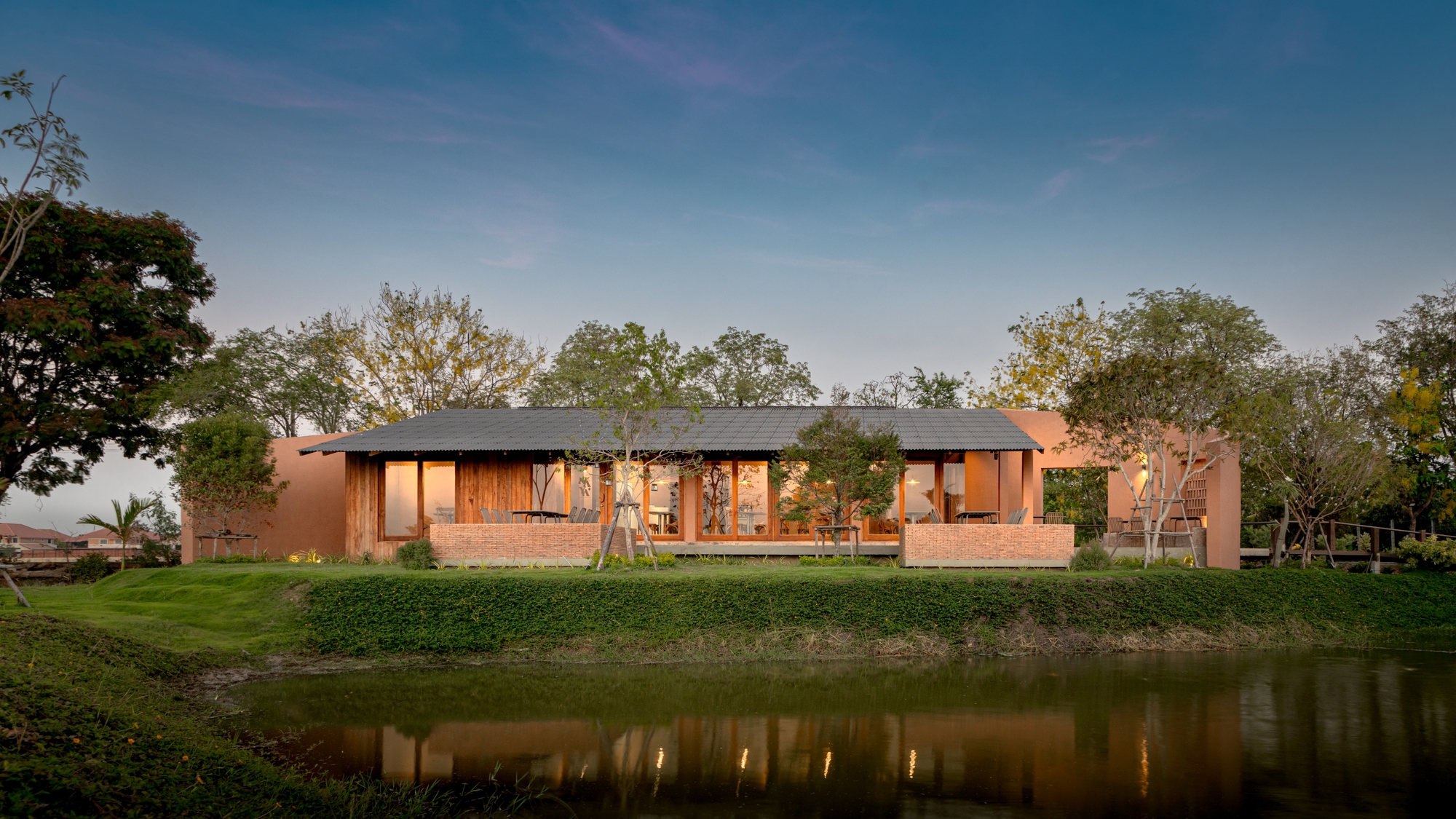 Illuminated timber and brick facade reflected in pond at twilight beneath a dark metal roof