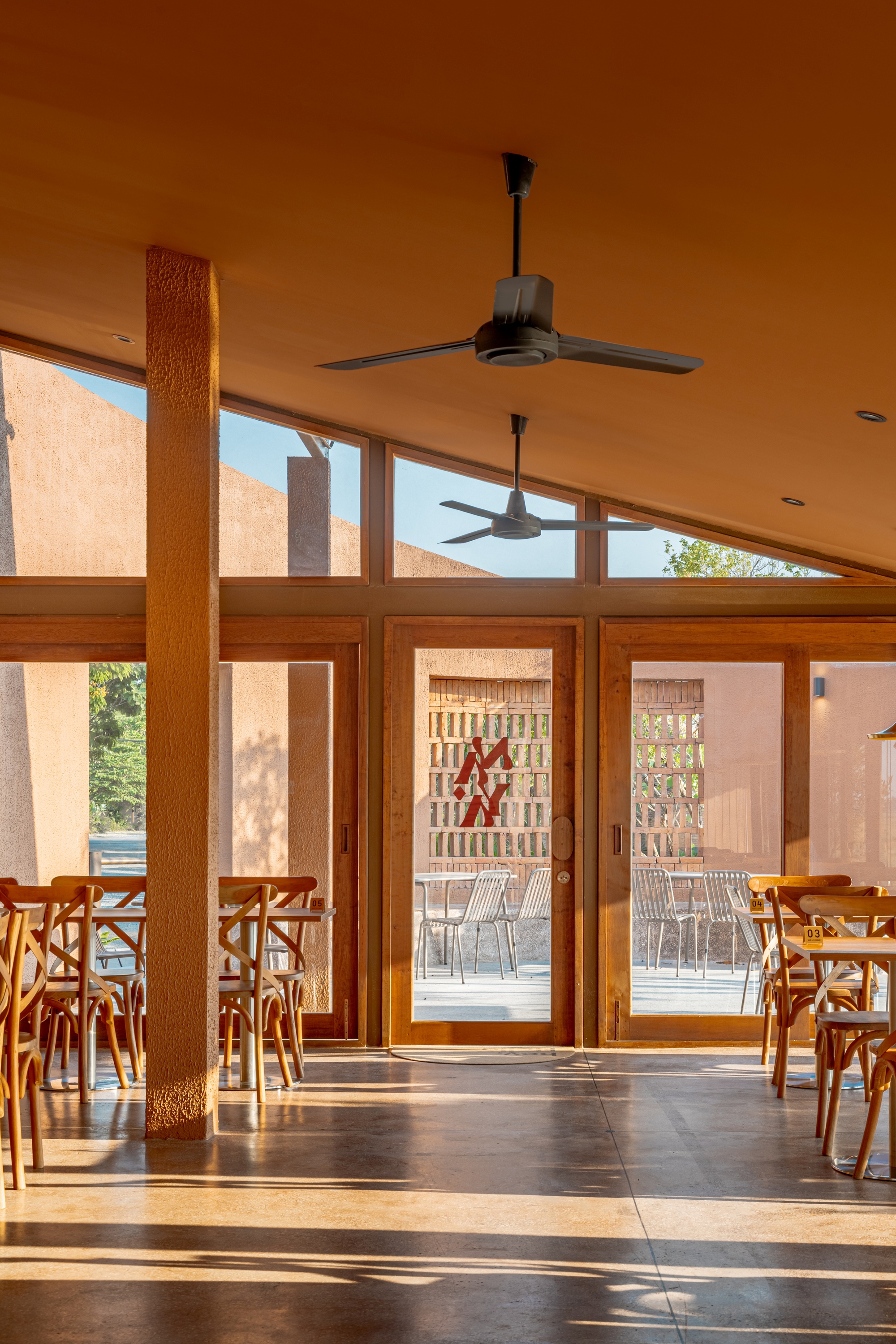 Interior dining space with timber-framed glazing and ceiling fans beneath a sloped ceiling in sunlight