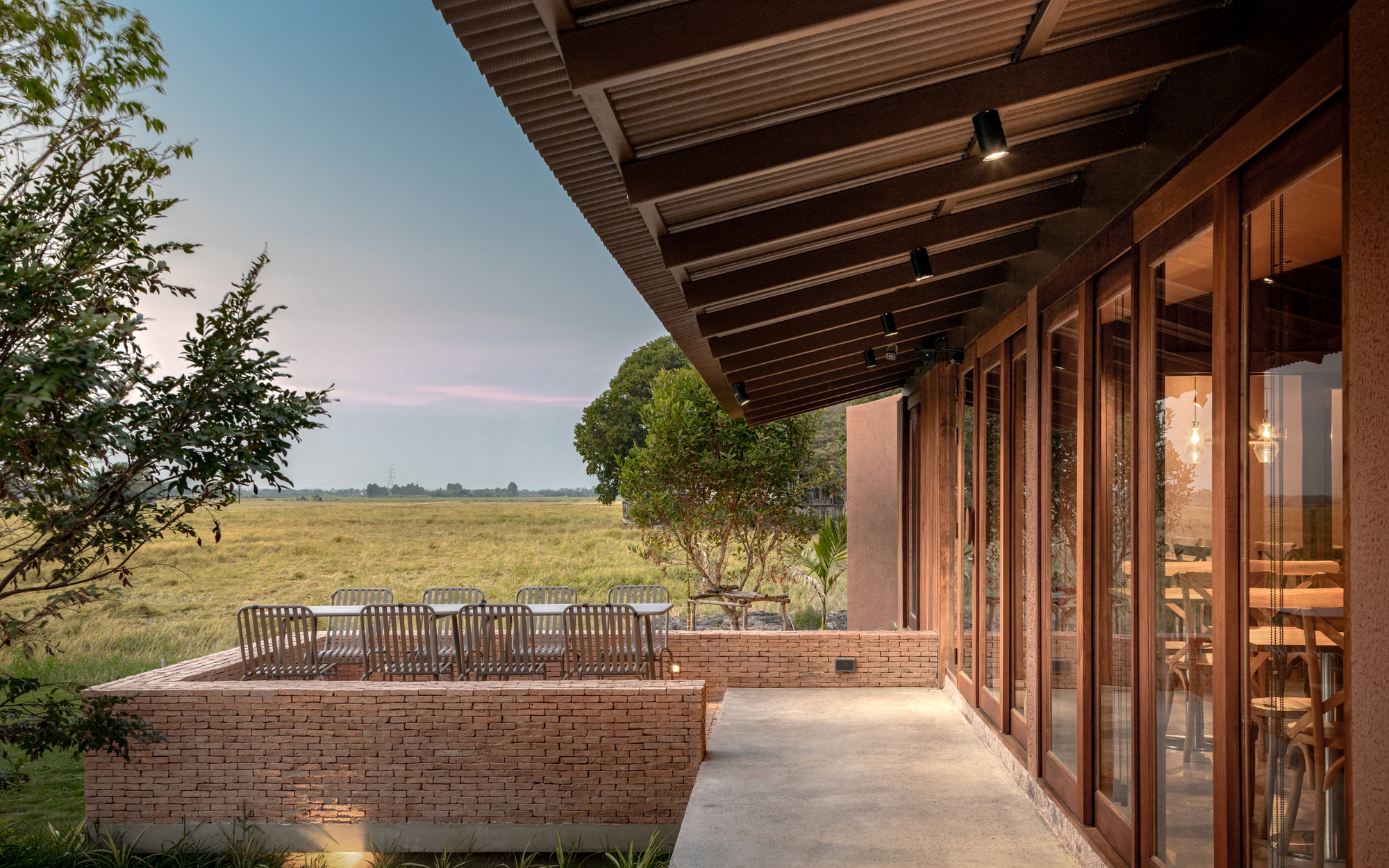 Deep overhang with exposed rafters sheltering timber-framed glazing and brick seating wall facing open fields