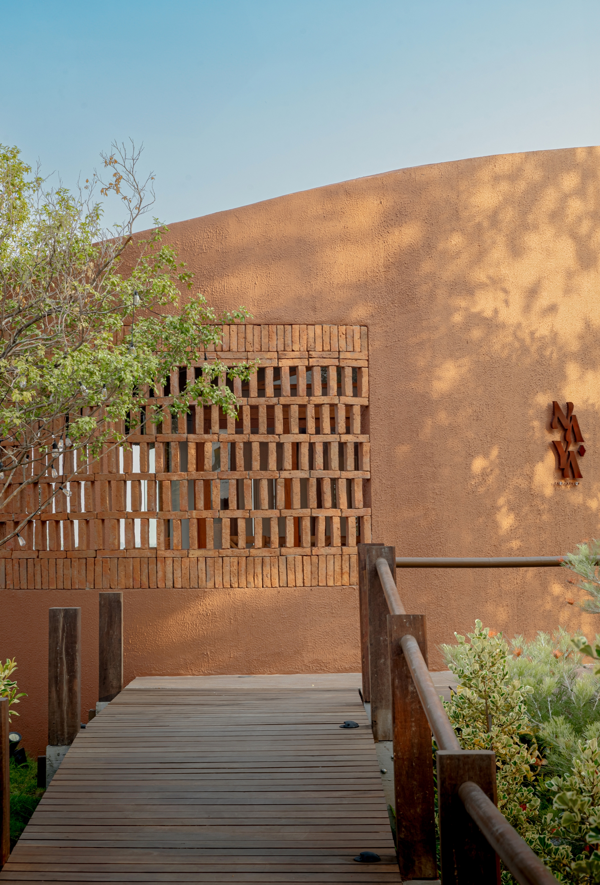 Textured exterior wall with lattice brick screen and timber bridge entry casting tree shadows