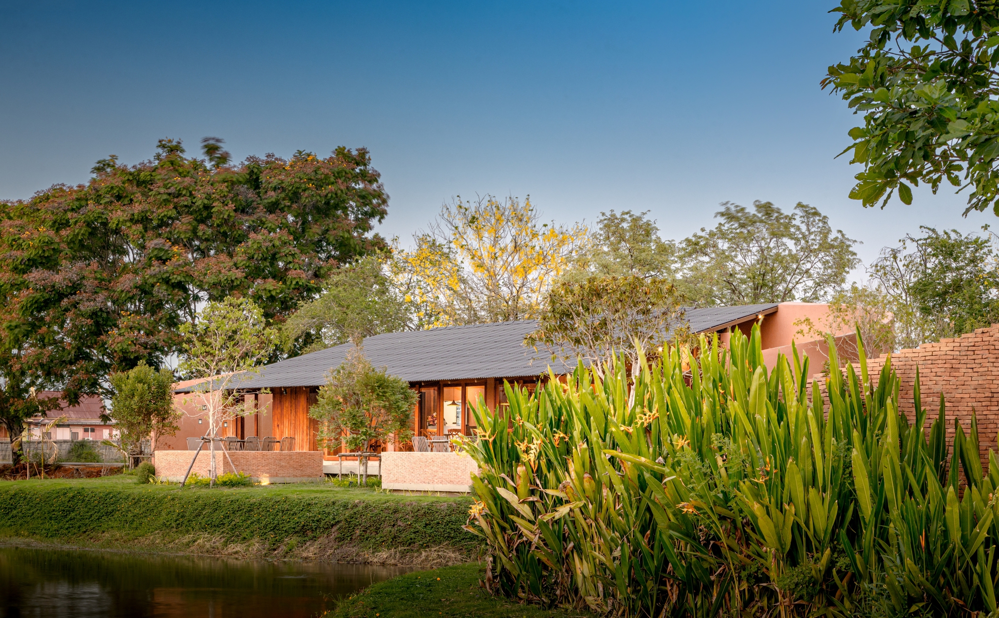 Low-slung pavilion with corrugated metal roof beside a pond edged with ornamental grasses at twilight