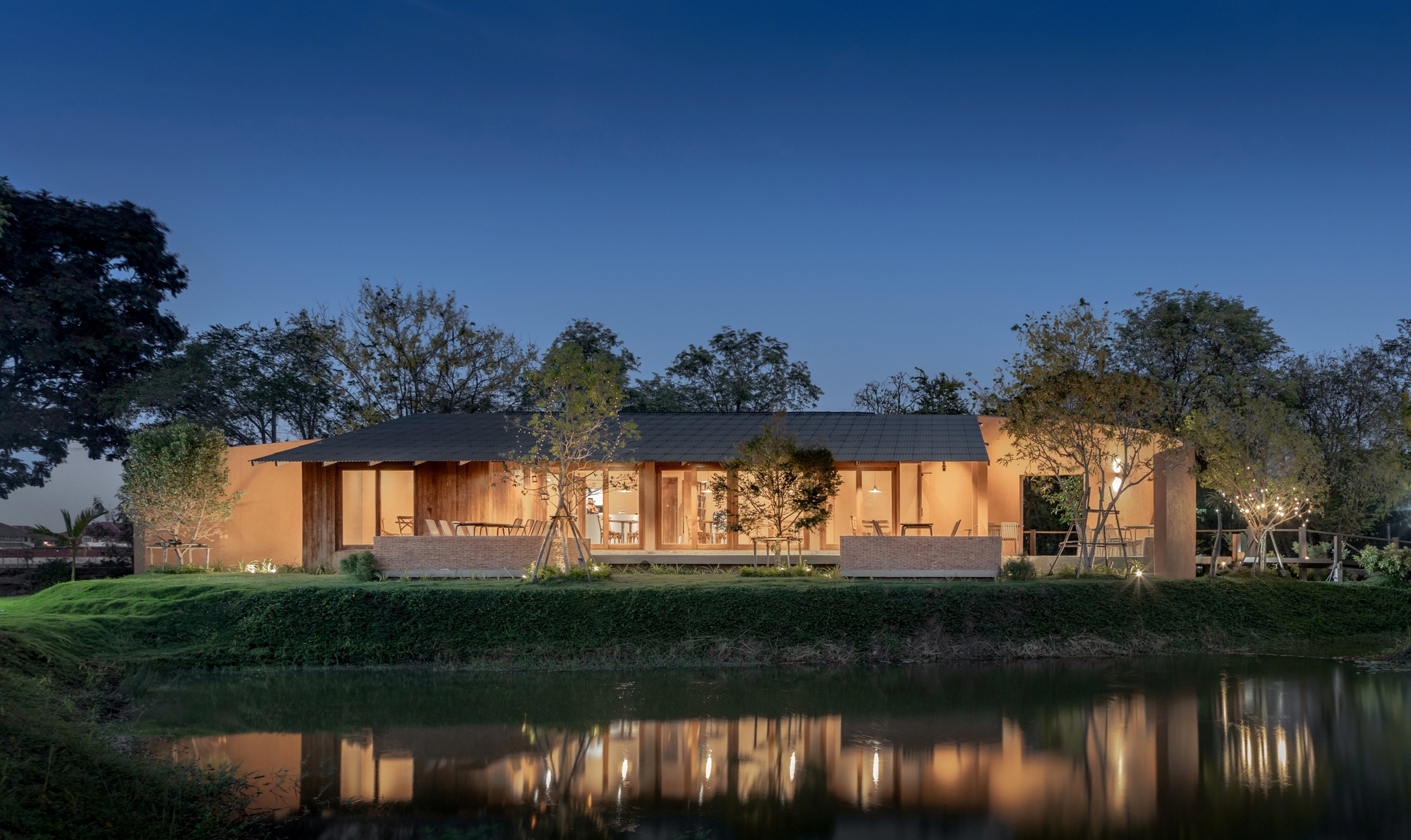 Illuminated single-story pavilion with glass walls reflected in a pond at dusk