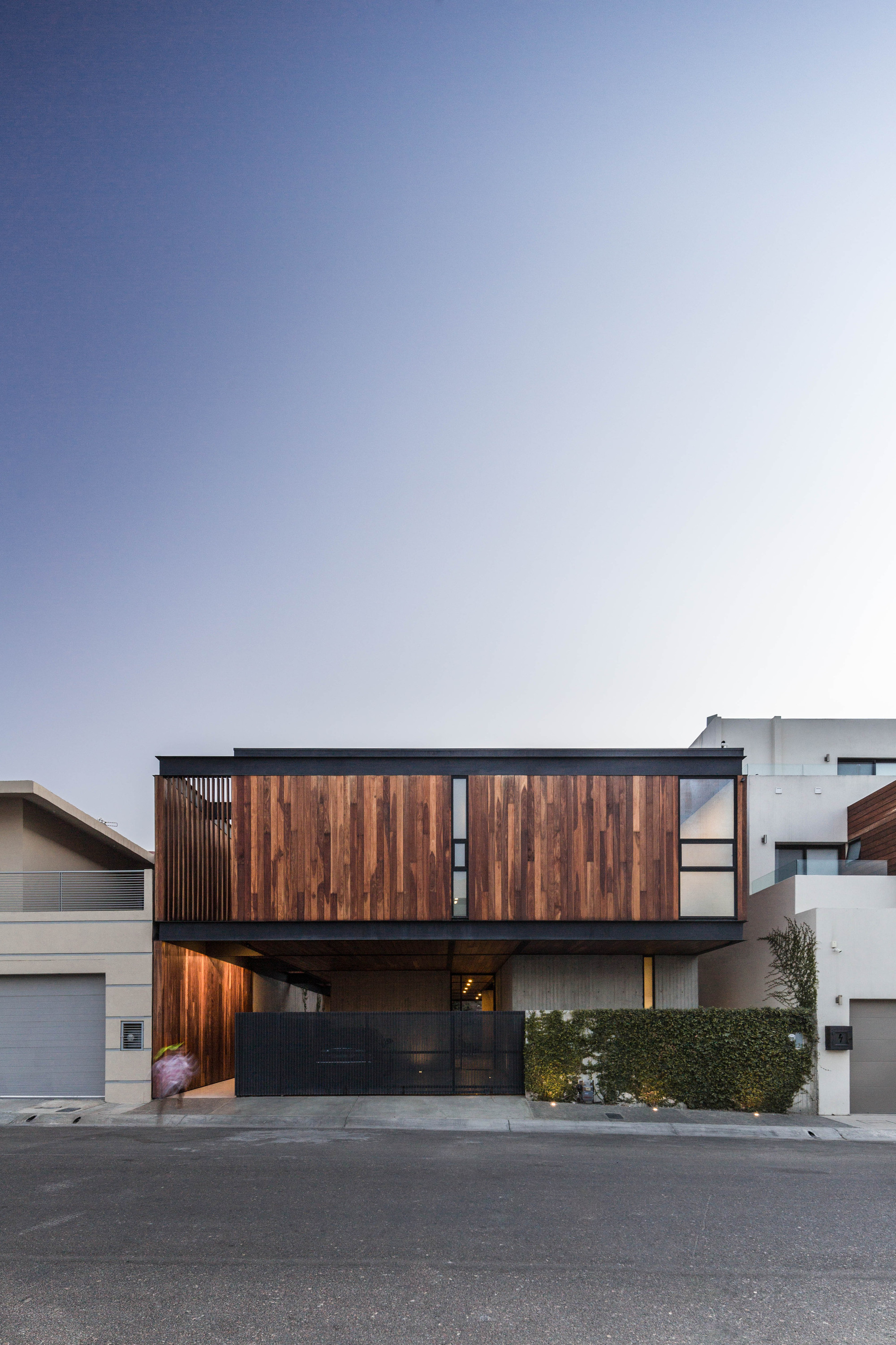 Street facade with timber-clad upper floor over a black steel gate and hedge at dusk