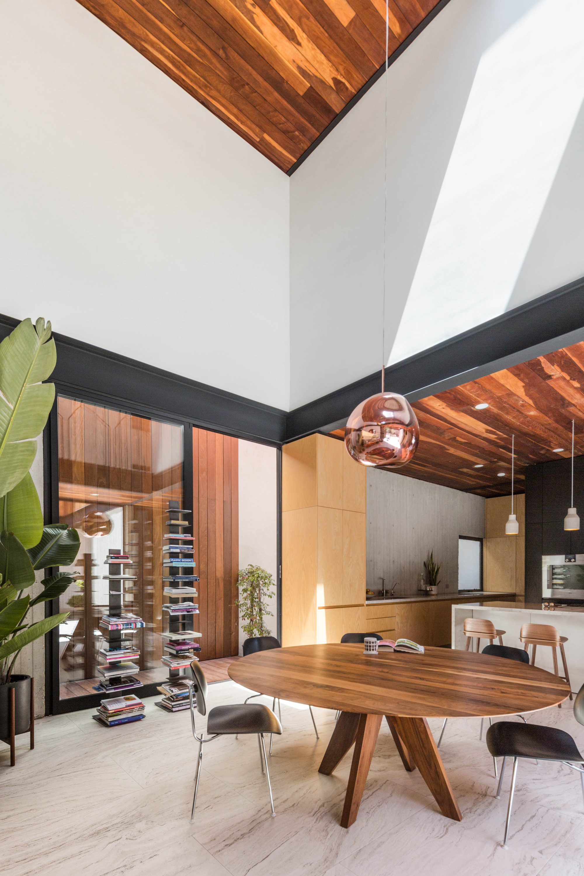 Double-height dining and kitchen area with a round wood table, copper pendant and sunlight on white walls beneath a timber ceiling
