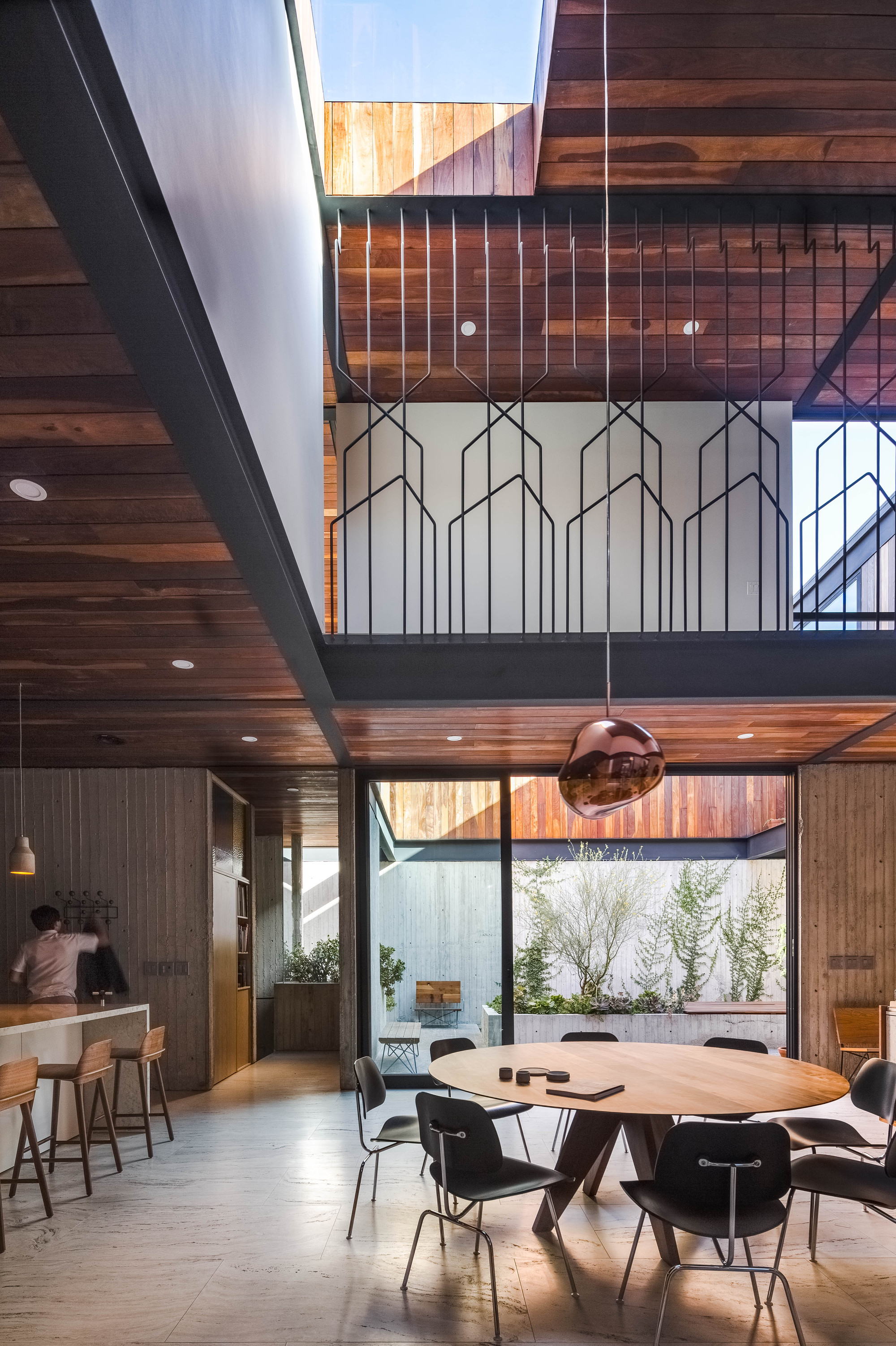 Double-height dining space with a round timber table, copper pendant lamp and patterned steel balustrade, lit by daylight from a courtyard