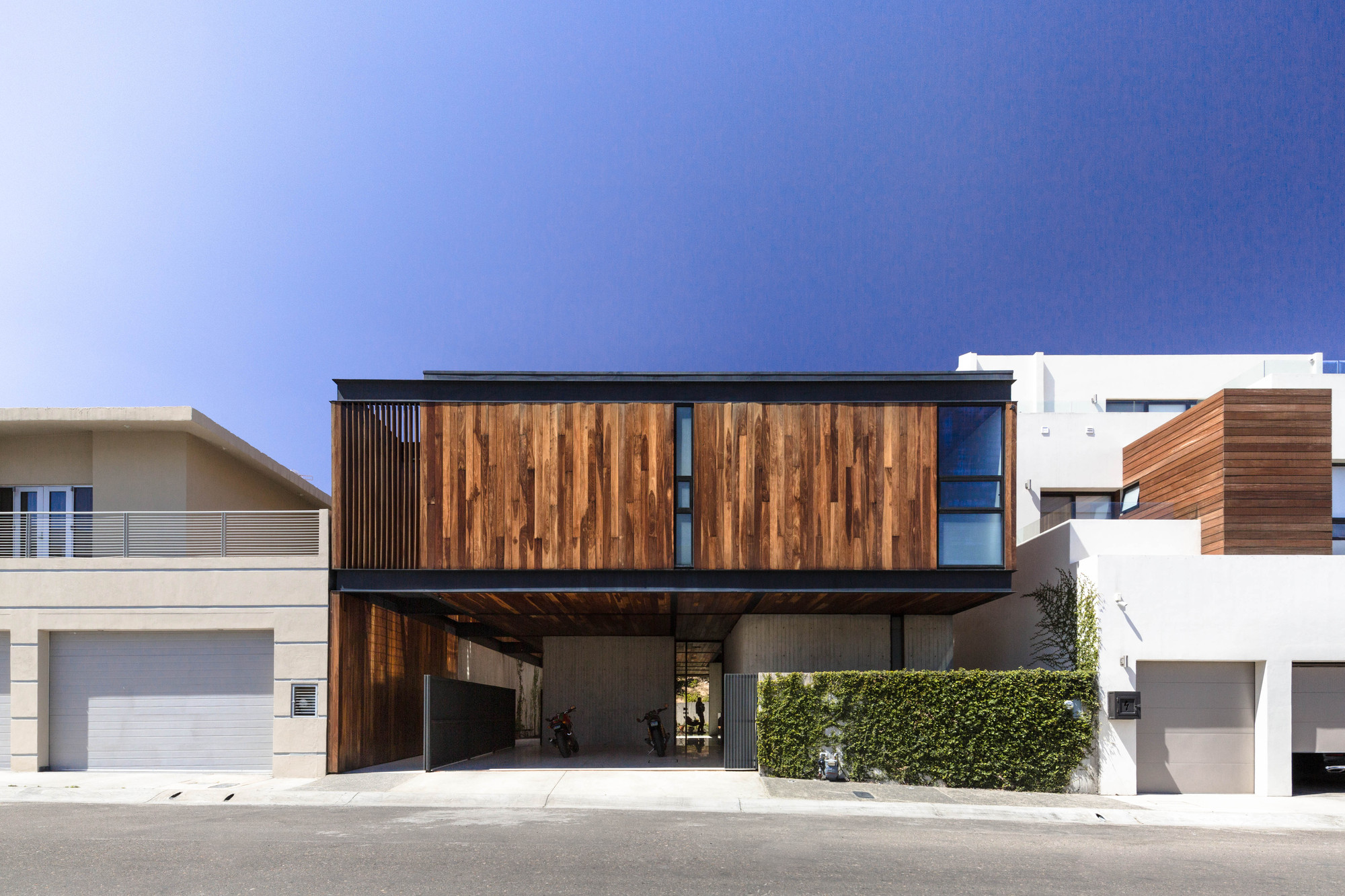 Street facade with a timber-clad upper volume floating above an open carport and a hedge, under a deep blue clear sky