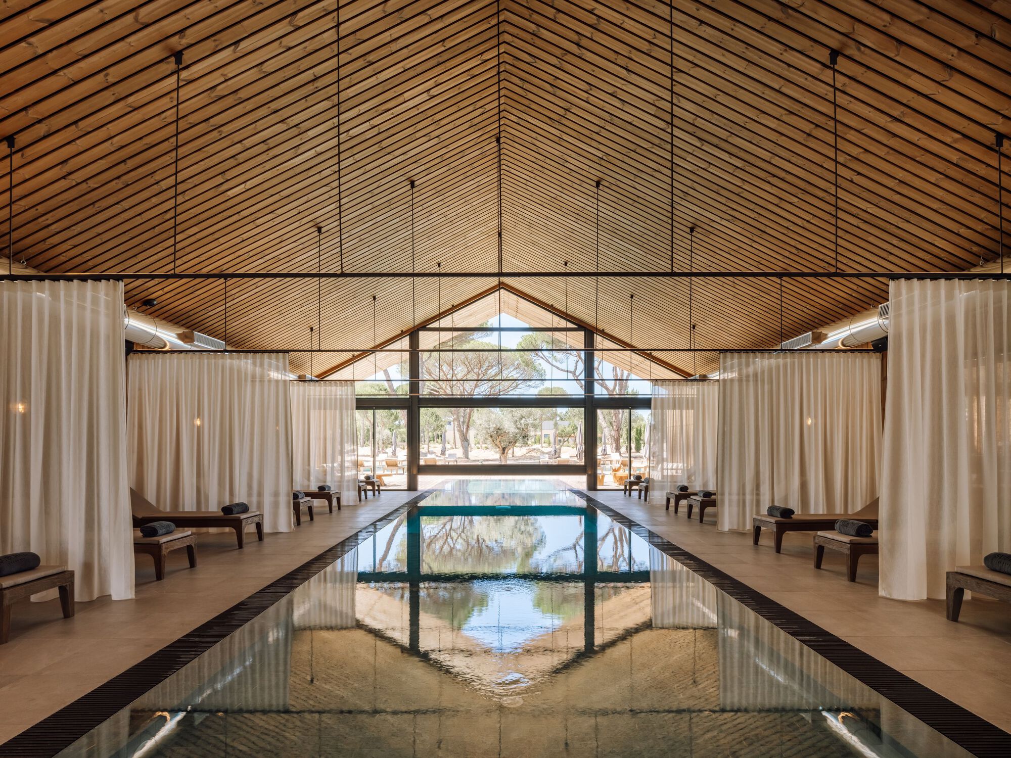 Interior lap pool under a gabled wood slat ceiling with white curtains flanking the lounging alcoves