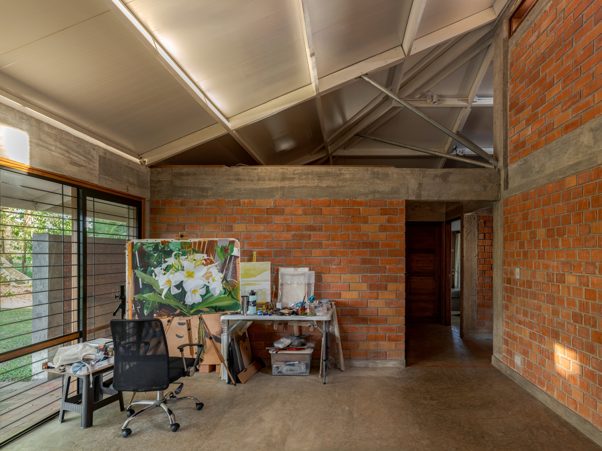 Interior studio space with exposed steel roof trusses, brick walls and an easel beside horizontal windows