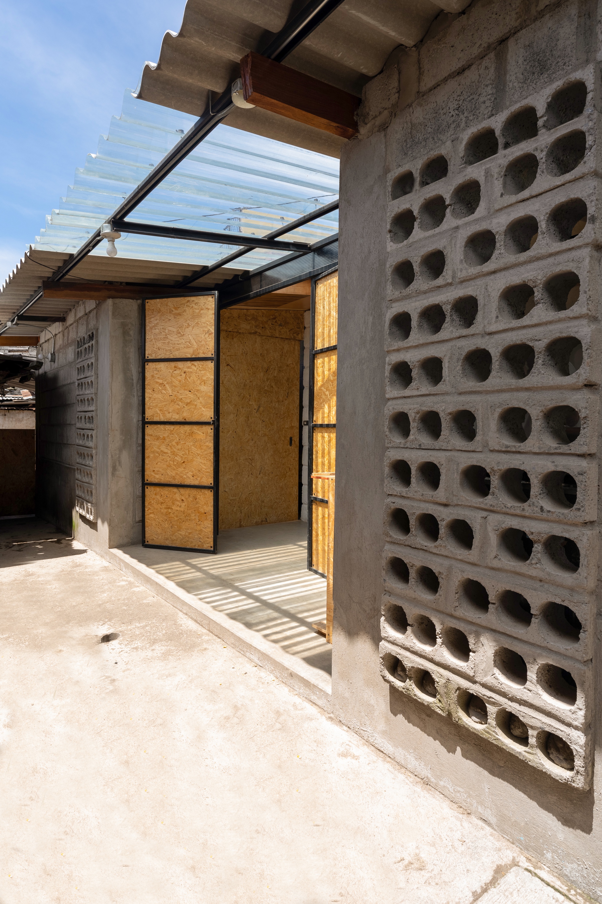 Exterior entryway with perforated concrete block wall and folding OSB doors beneath a translucent canopy