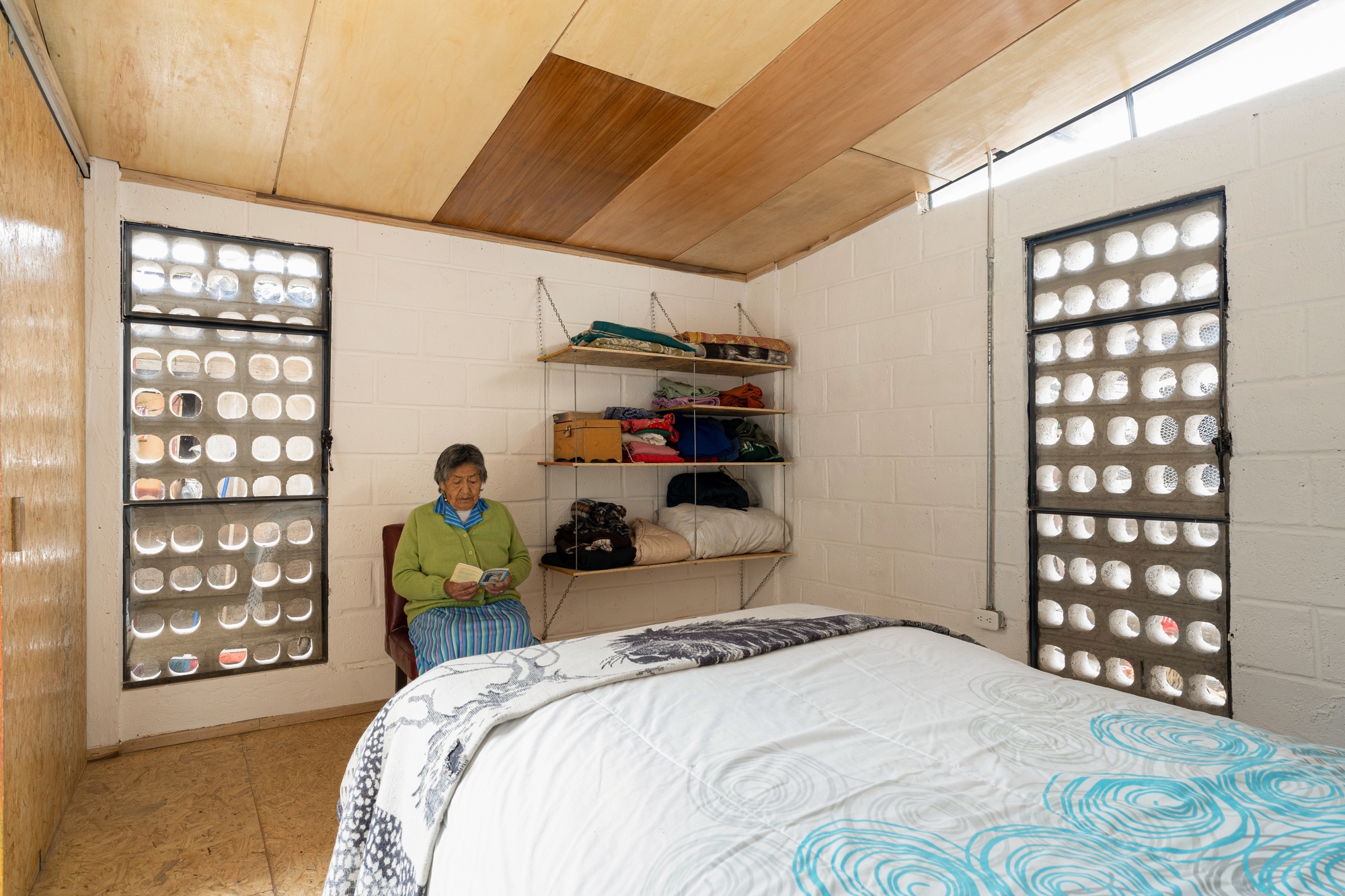 Bedroom with concrete block walls and two glass block windows flanking a resident on a chair