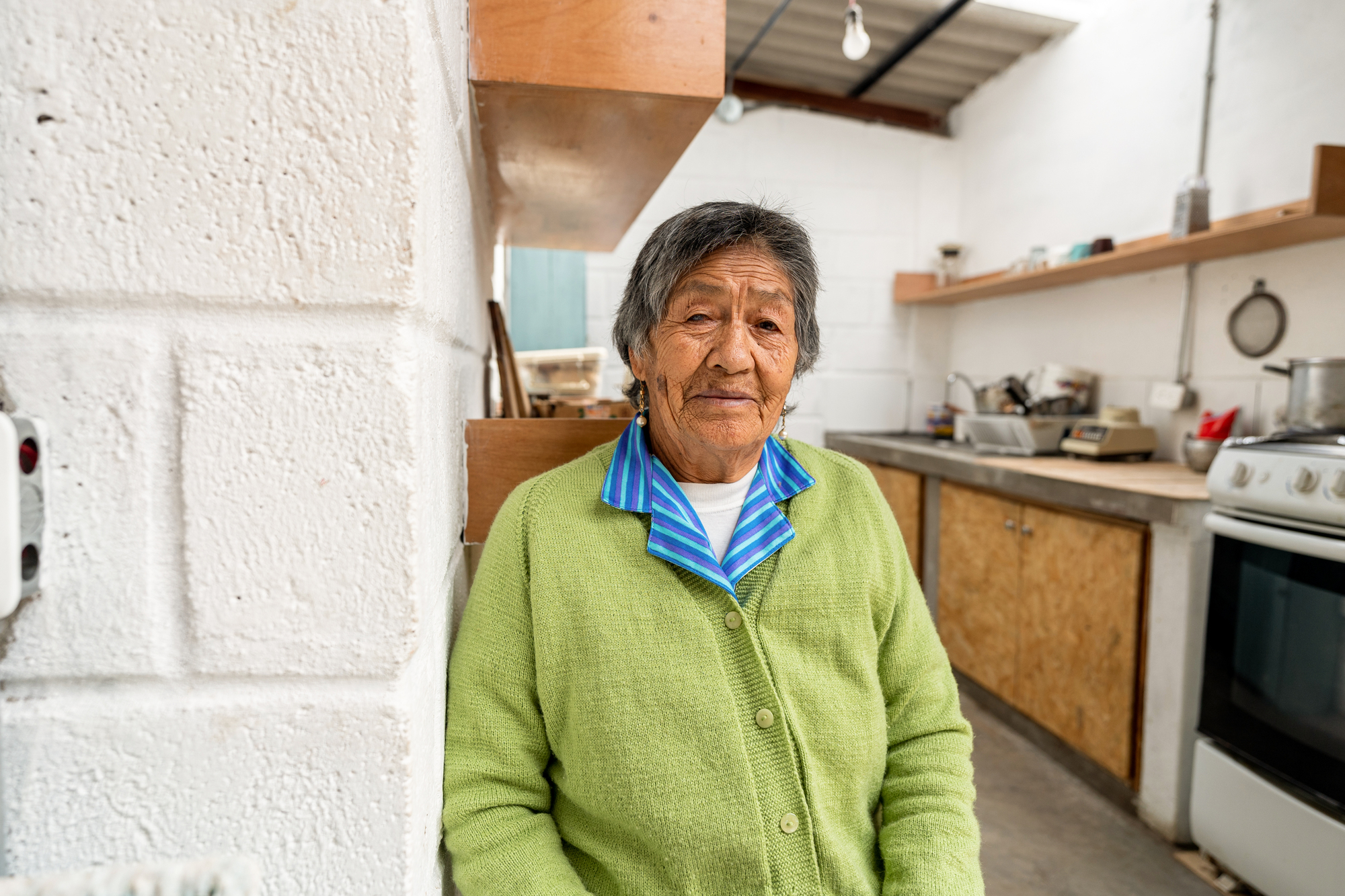 Resident standing in kitchen with plywood cabinetry, open shelving, and whitewashed masonry walls under exposed timber rafters