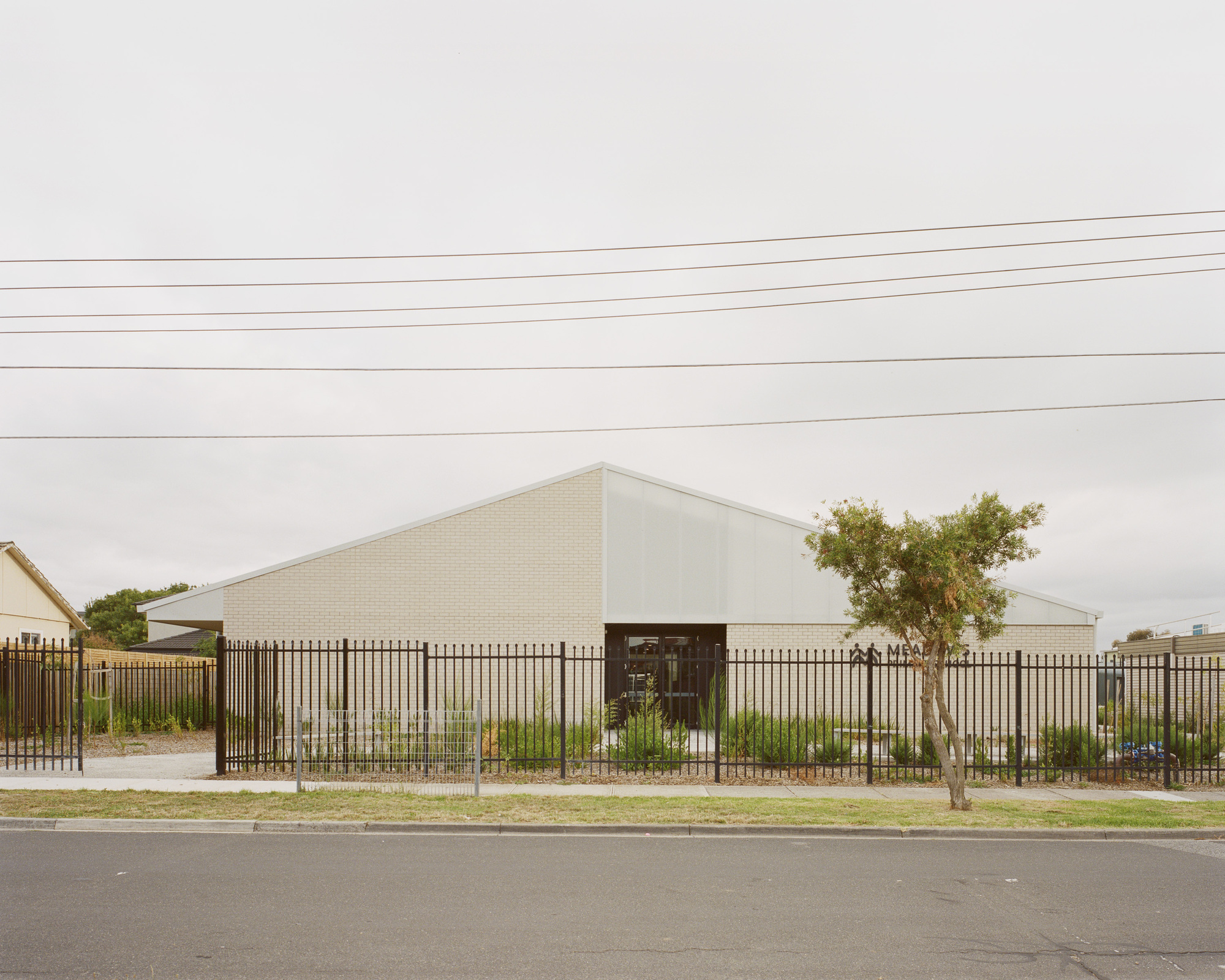 Street view of the white brick gabled volume behind vertical steel fencing and young trees