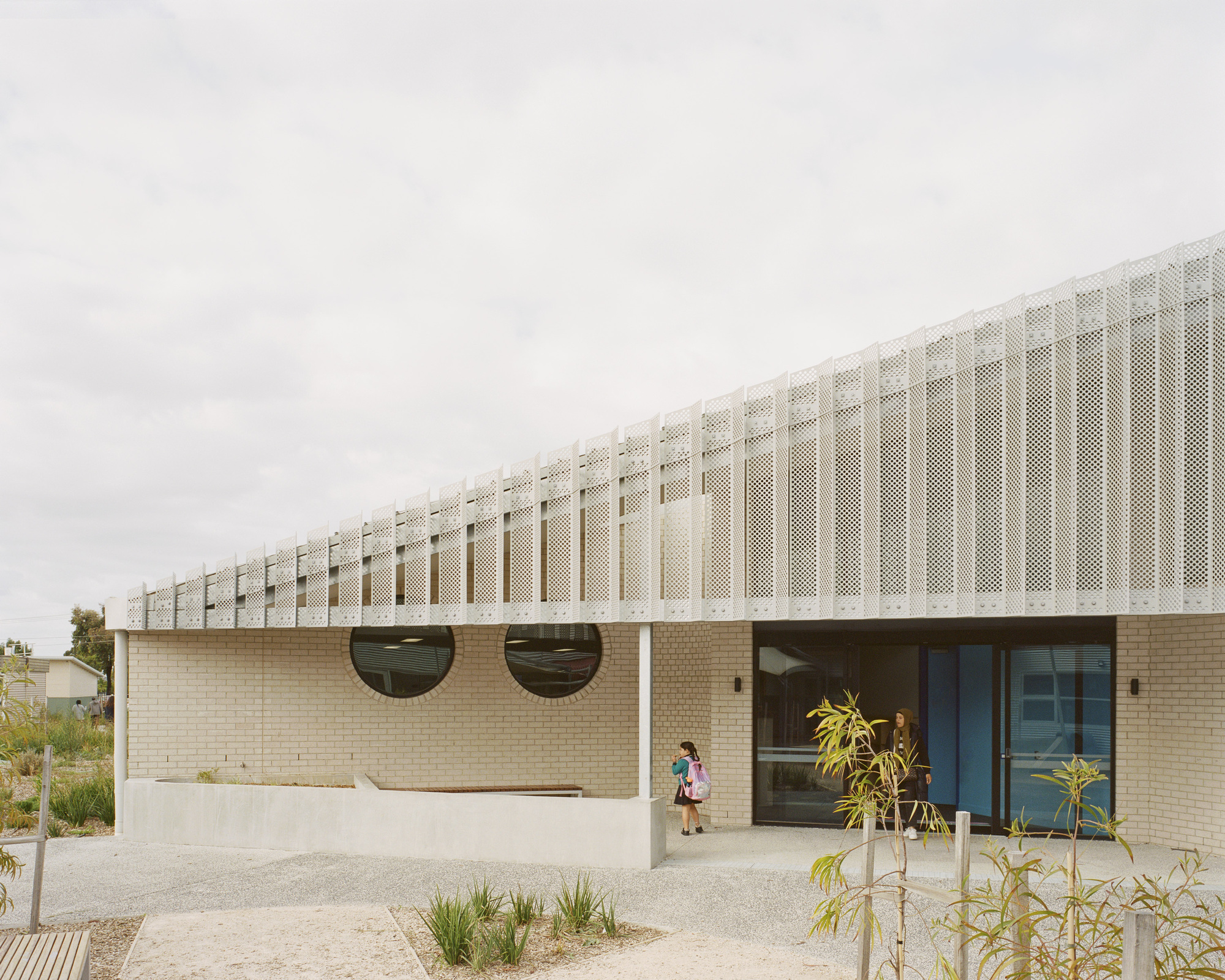 Perforated metal screen above cream brick base with circular windows and a child at the entrance