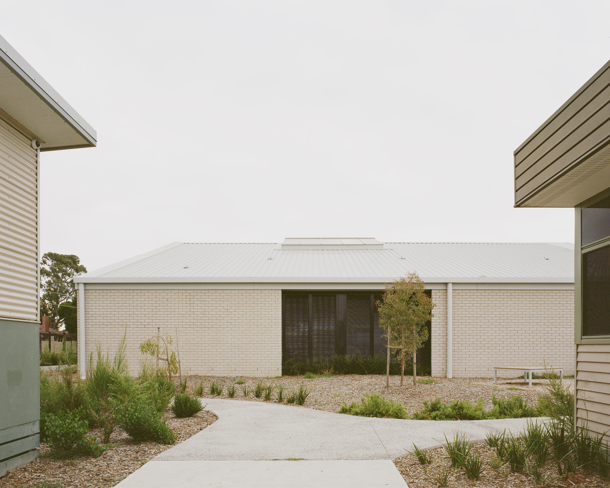 Central courtyard framed by corrugated metal volumes and a low cream brick pavilion under overcast sky